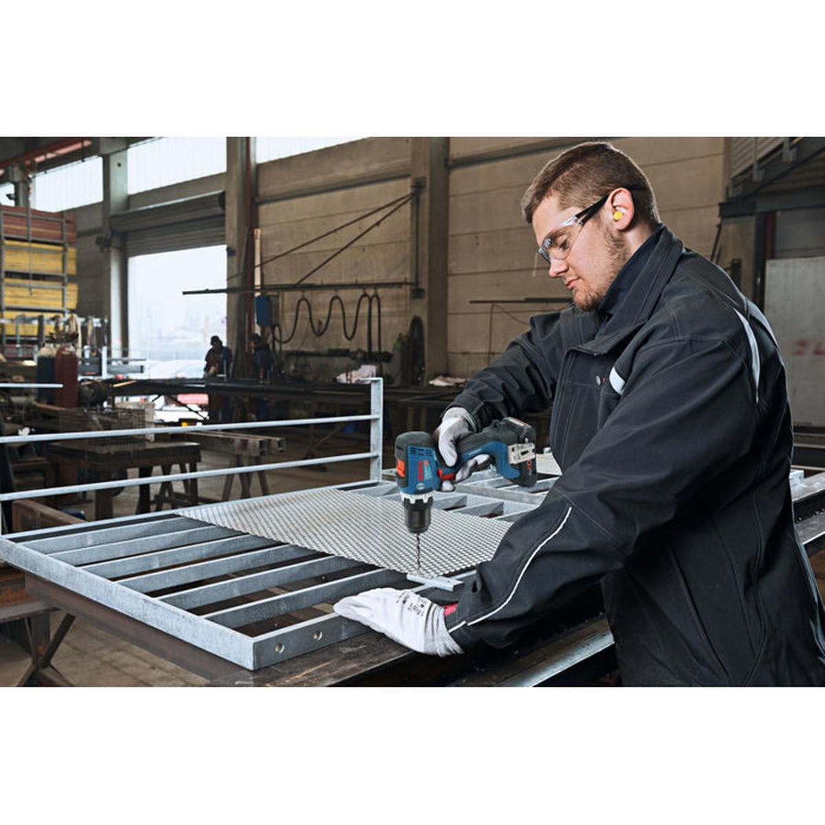 Ein Arbeiter in Schutzbrille bohrt mit einem Akku-Schrauber in eine Metallkonstruktion in einer Werkstatt.