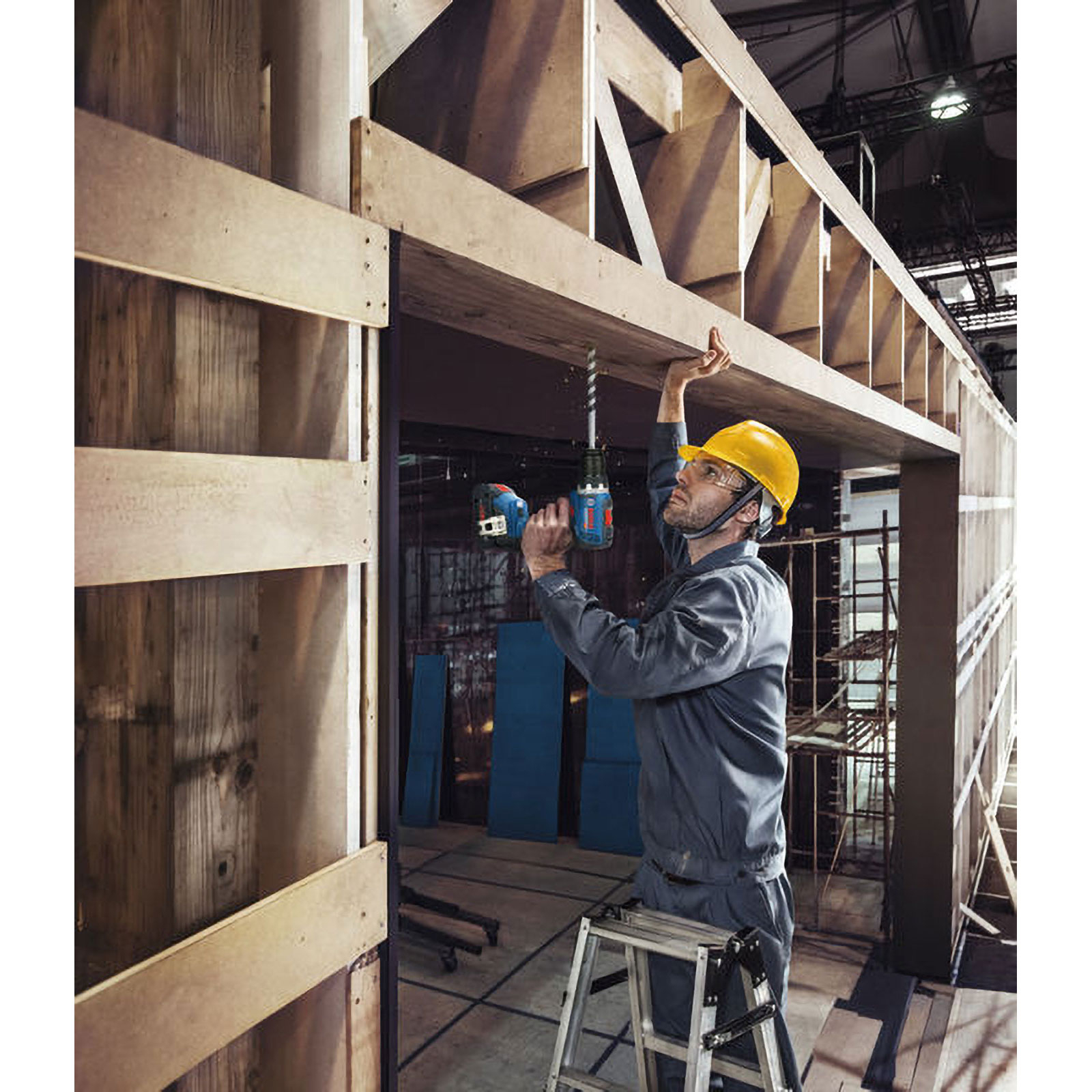 Ein Arbeiter in gelbem Helm steht auf einer Leiter und bohrt in eine Holzstruktur.