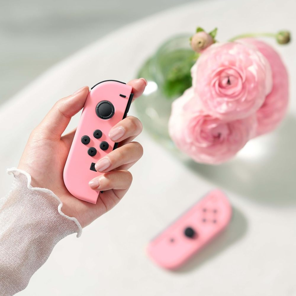 Eine Hand hält einen rosa Controller, mit rosa Blumen und einem weiteren Controller im verschwommenen Hintergrund.