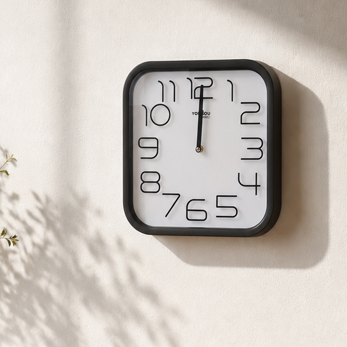 Eine quadratische Wanduhr mit schwarzem Rahmen und weißem Zifferblatt hängt an einer beigen Wand mit Pflanzenschatten.