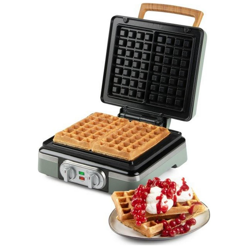 Una piastra per waffle verde aperta contiene due waffle dorati, con un altro waffle su un piatto guarnito di crema e bacche rosse.