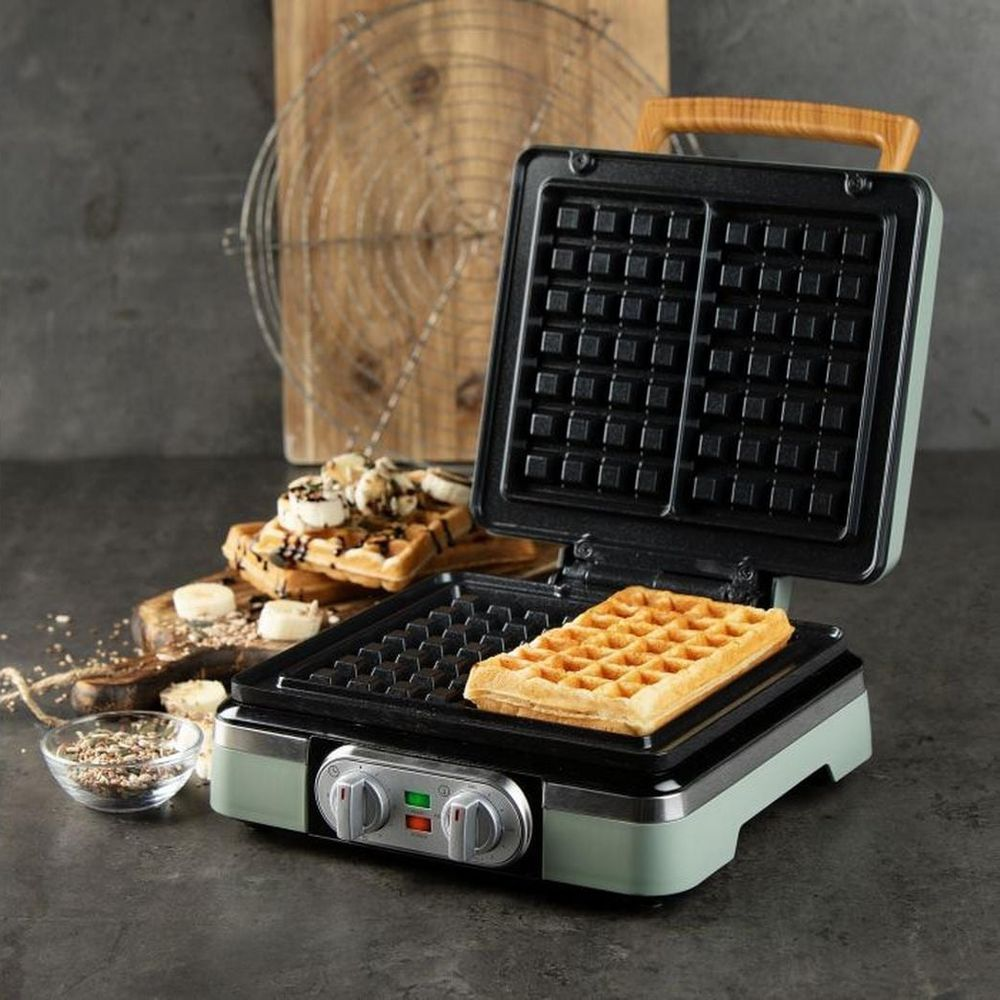 Una cialdiera verde chiaro cuoce un waffle dorato, con waffle a banana e cioccolato sullo sfondo.
