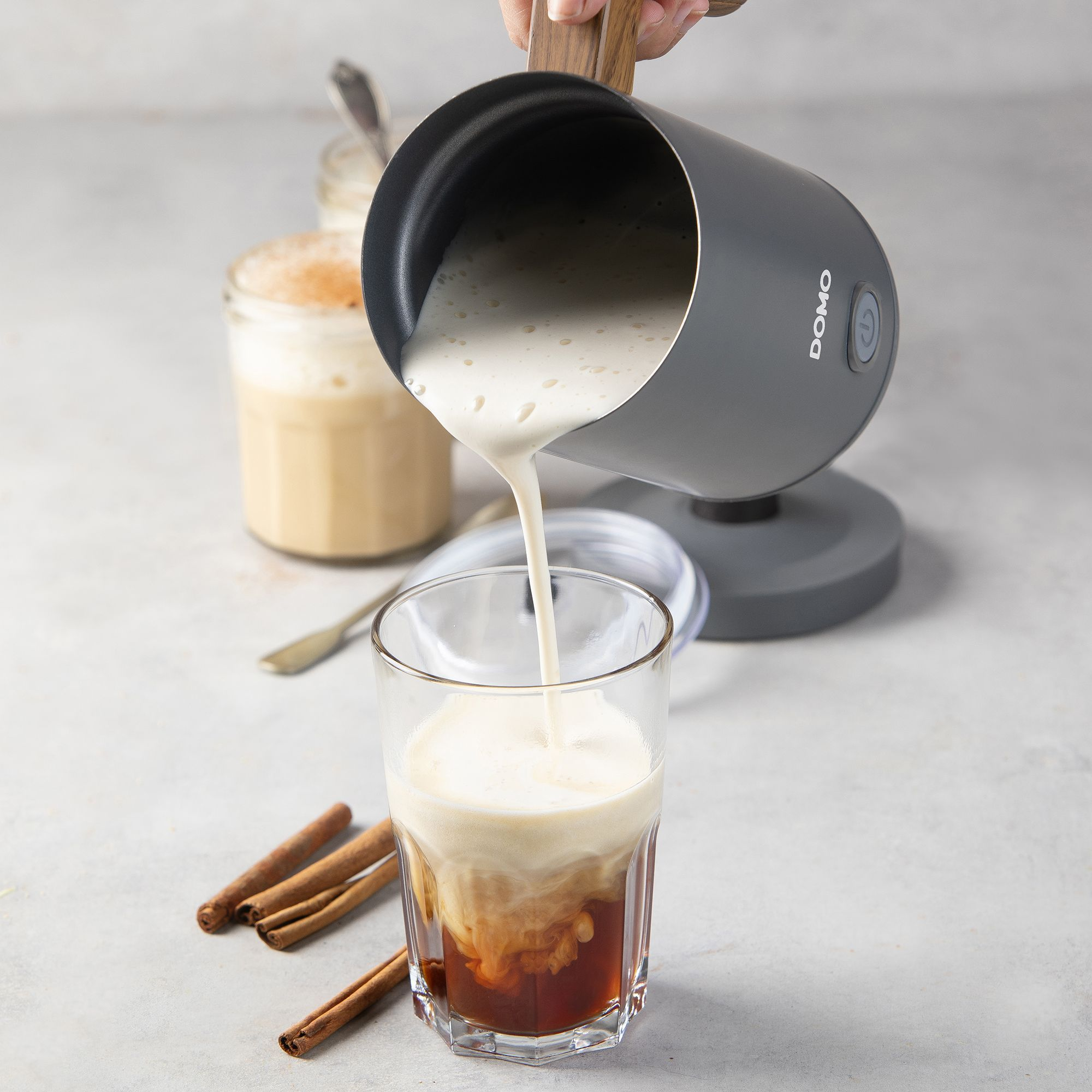 Milchschaum wird aus einer grauen Kanne in ein Glas mit Kaffee gegossen, nebeneinander Zimtstangen.
