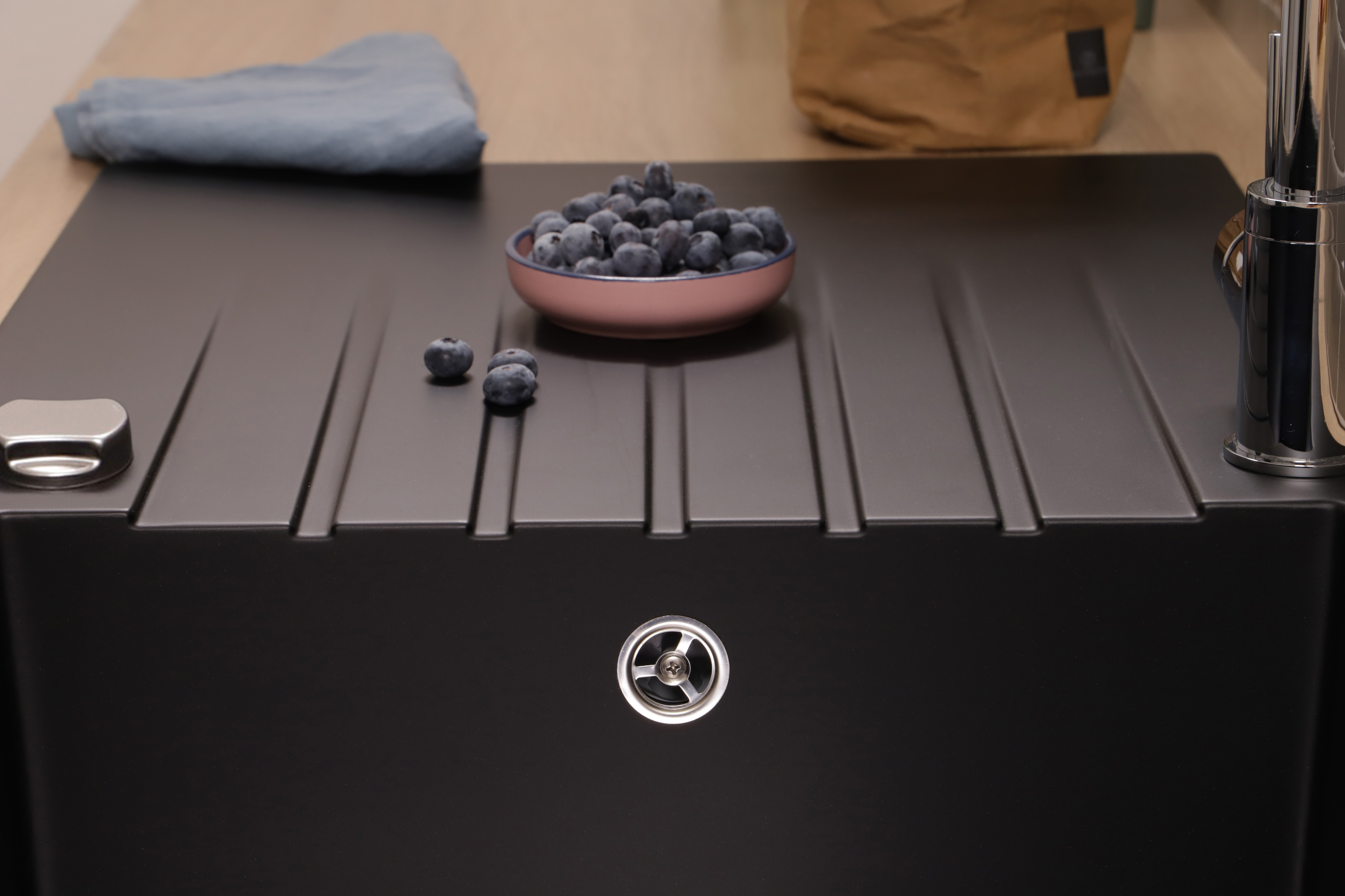 Blaubeeren in einer Schüssel und drei lose Blaubeeren auf einer schwarzen Spüle, daneben ein graues Tuch.