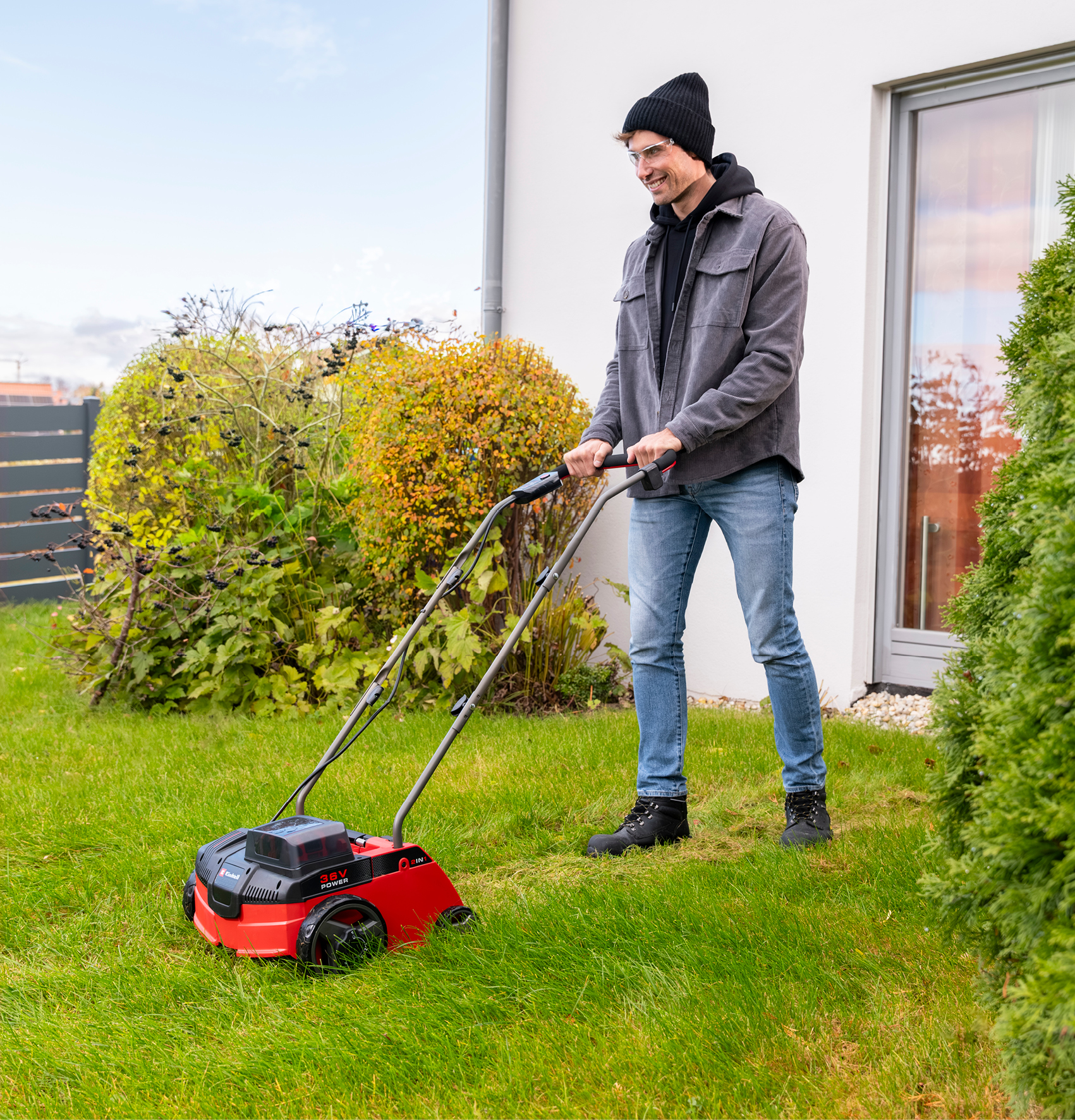 Ein lächelnder Mann in Mütze und grauer Jacke benutzt einen roten Rasenlüfter auf grünem Gras.