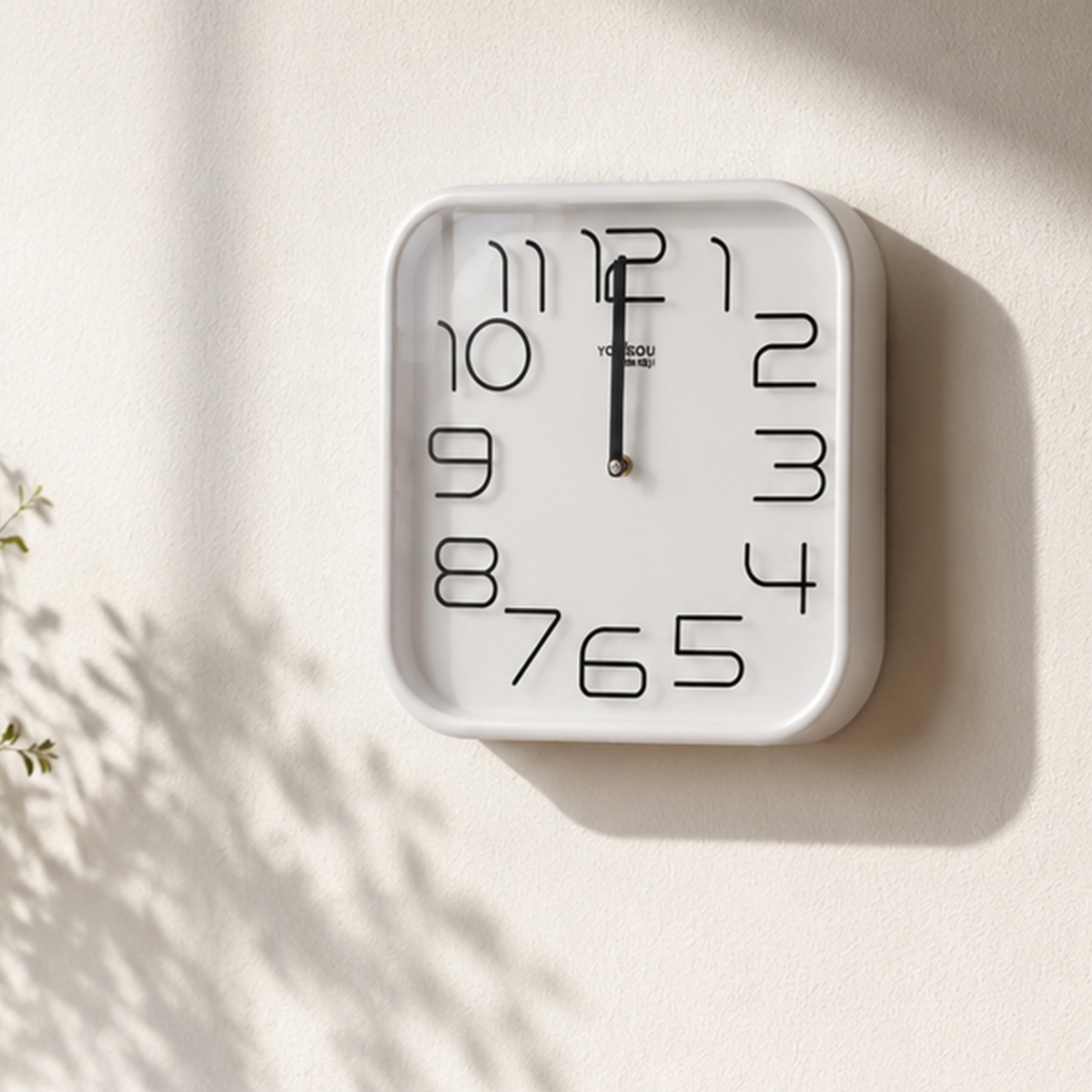 Eine quadratische weiße Wanduhr mit großen schwarzen Zahlen hängt an einer beigefarbenen Wand mit Schatten.