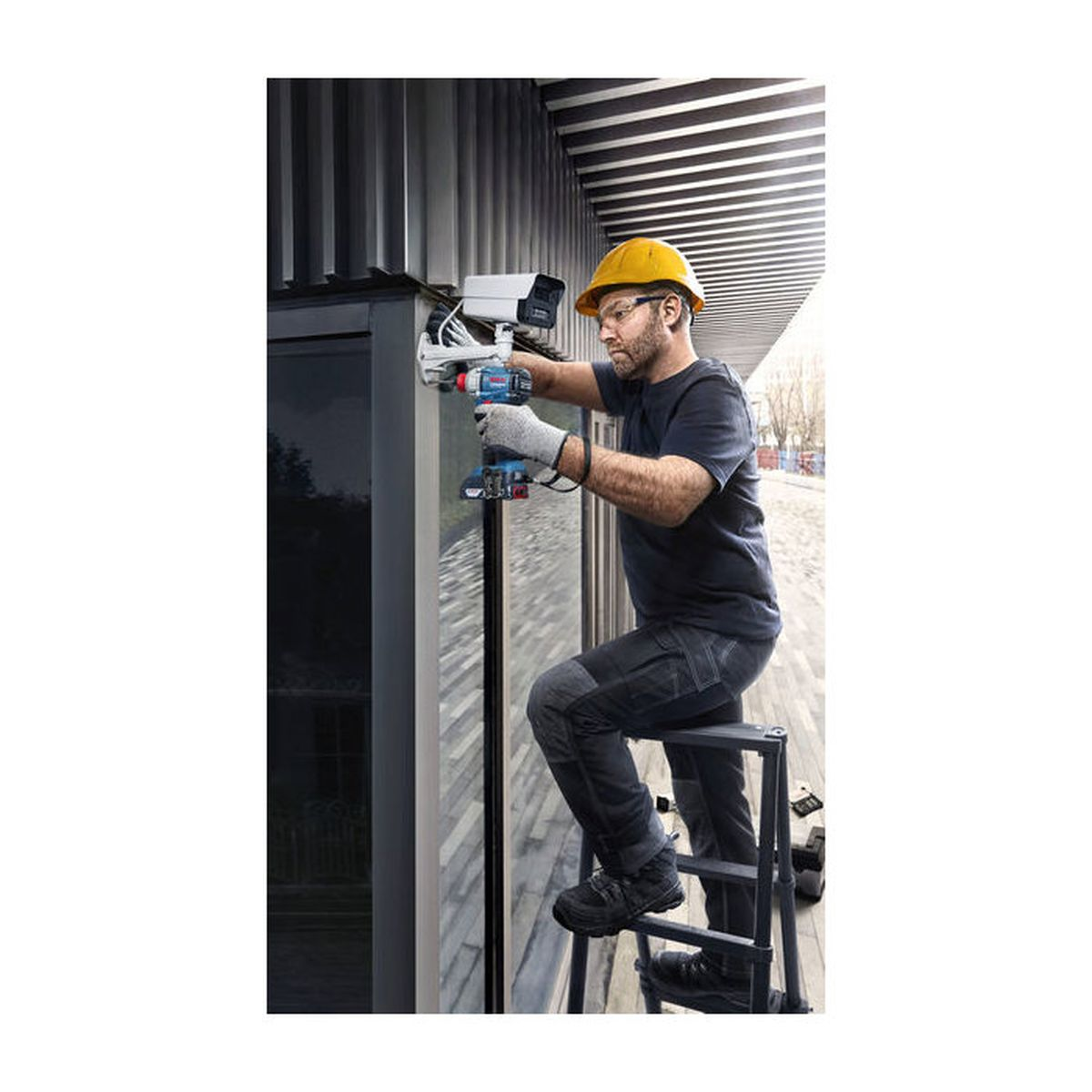 Ein Arbeiter mit gelbem Helm auf einer Leiter installiert mit einer Bohrmaschine eine Überwachungskamera.