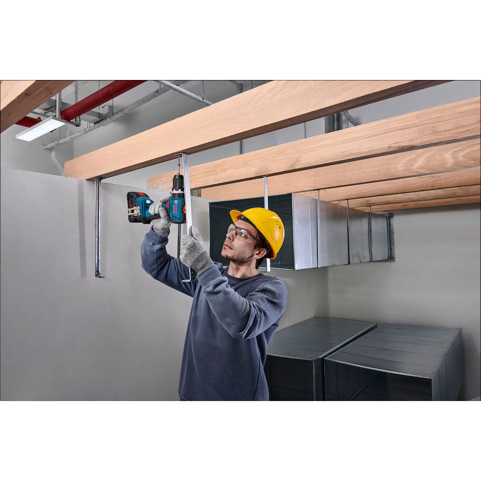 Ein Arbeiter mit gelbem Helm und Schutzbrille bohrt eine Metallhalterung an einen Deckenbalken aus Holz.