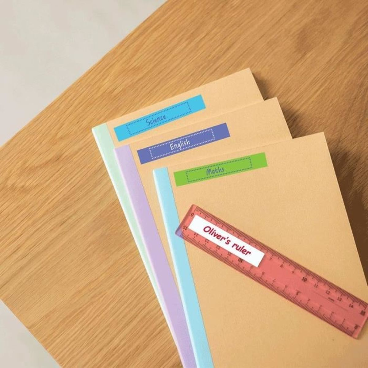 Drei braune Hefte mit farbigen Etiketten und ein Lineal liegen auf einem Holztisch.