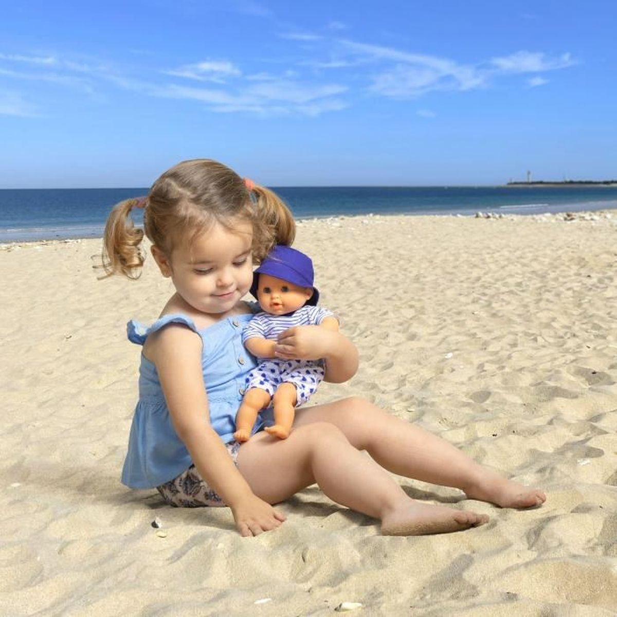 Kleines Mädchen mit Zöpfen sitzt am Strand und hält eine Puppe im Arm vor dem blauen Meer.