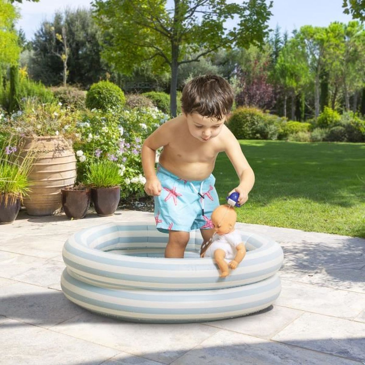 Kleiner Junge in Badehose steht in einem kleinen runden Planschbecken im Garten und wäscht eine Puppe.