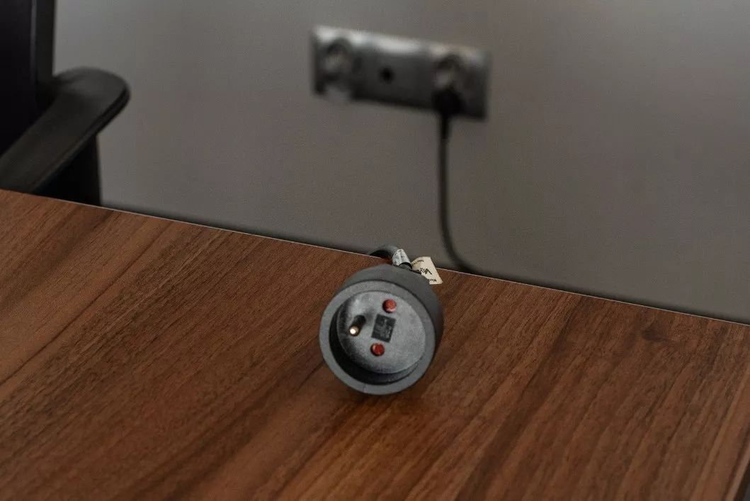 Ein schwarzer Stecker liegt auf einem Holztisch, das Kabel führt zu einer Wandsteckdose.