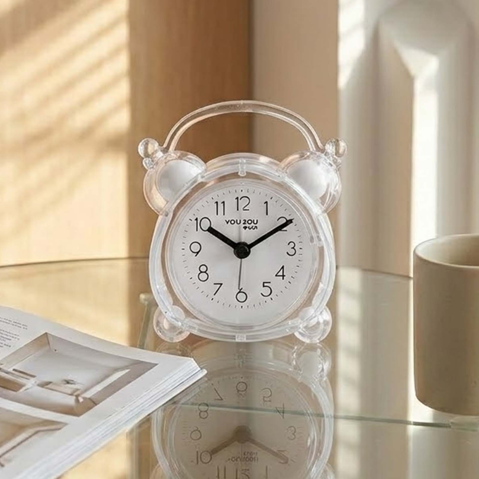Pequeño reloj despertador transparente en mesa de cristal con libro y taza.