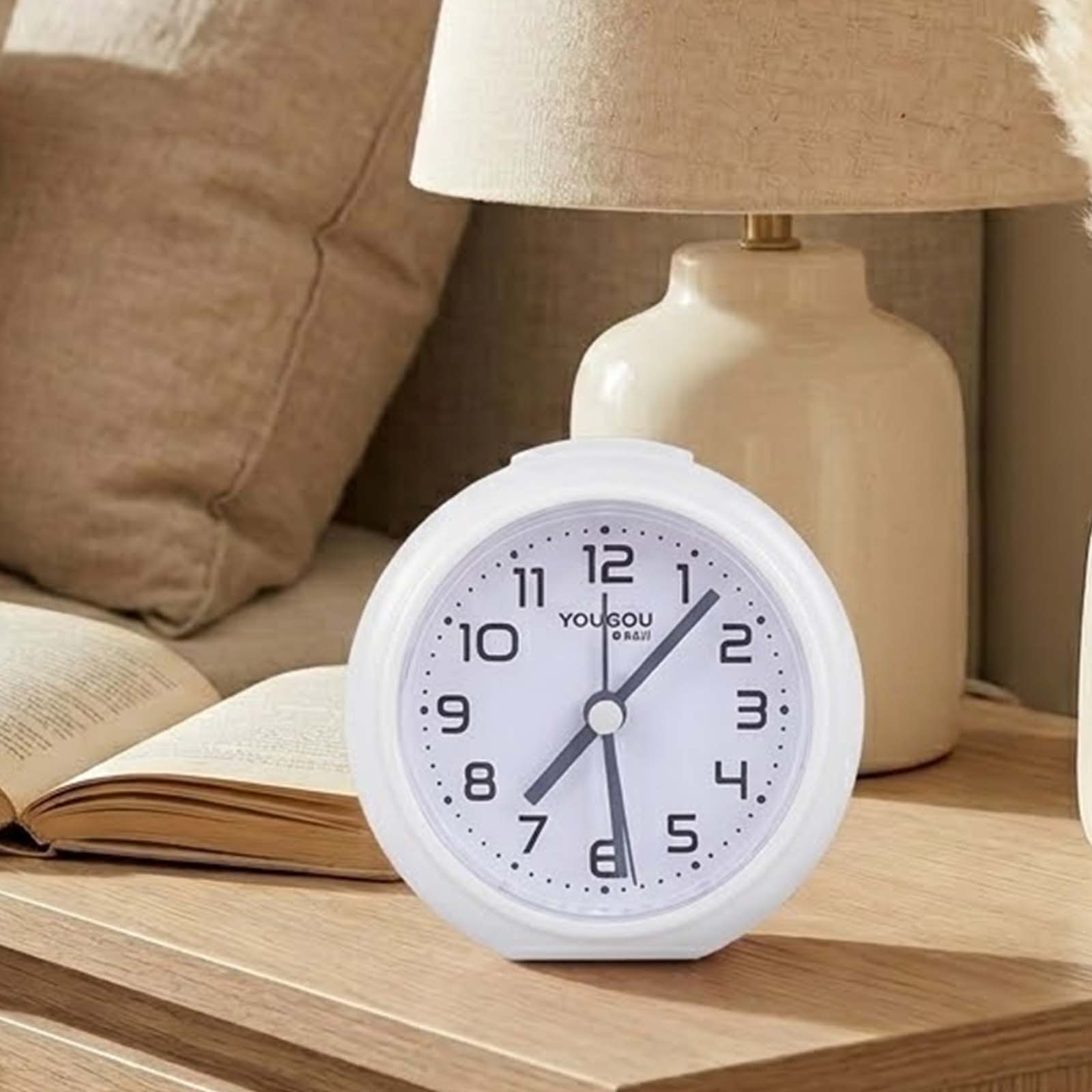 Un despertador analógico blanco sobre una mesa de madera, con un libro abierto, una lámpara y un cojín.