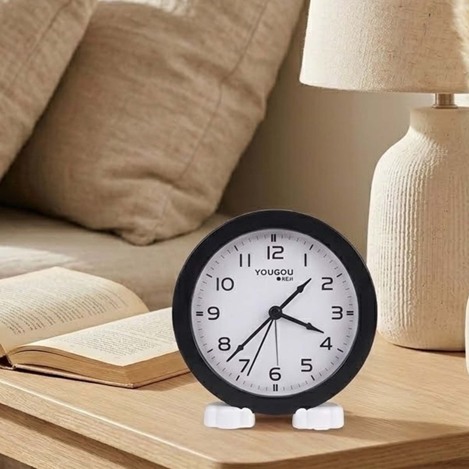 Un reloj de mesa negro con esfera blanca sobre una mesa de madera, al lado de un libro abierto y una lámpara.