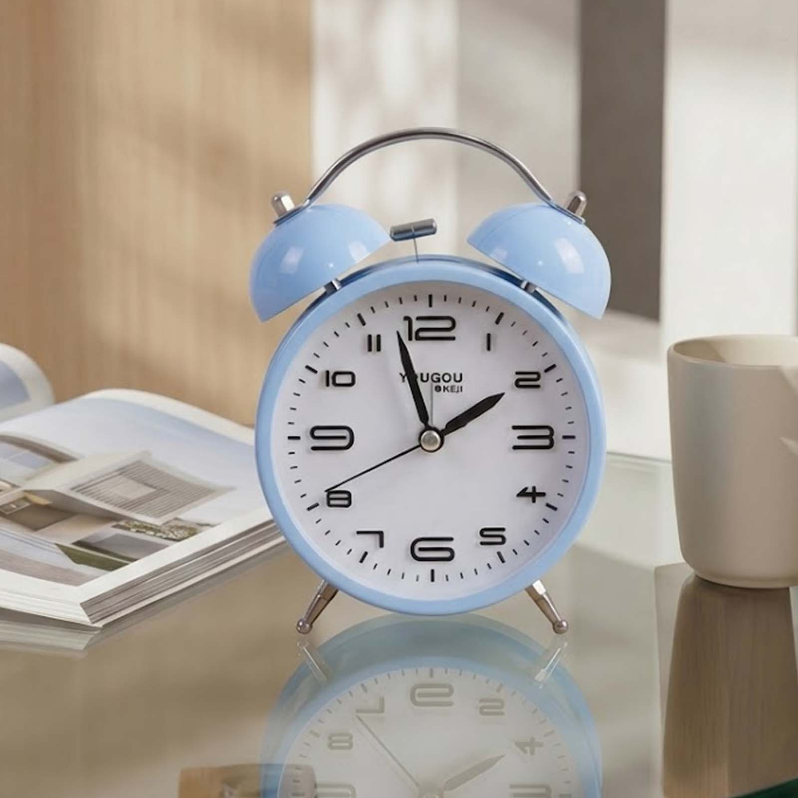 Un despertador azul claro con doble campana se encuentra en una mesa de cristal junto a una revista abierta y una taza.