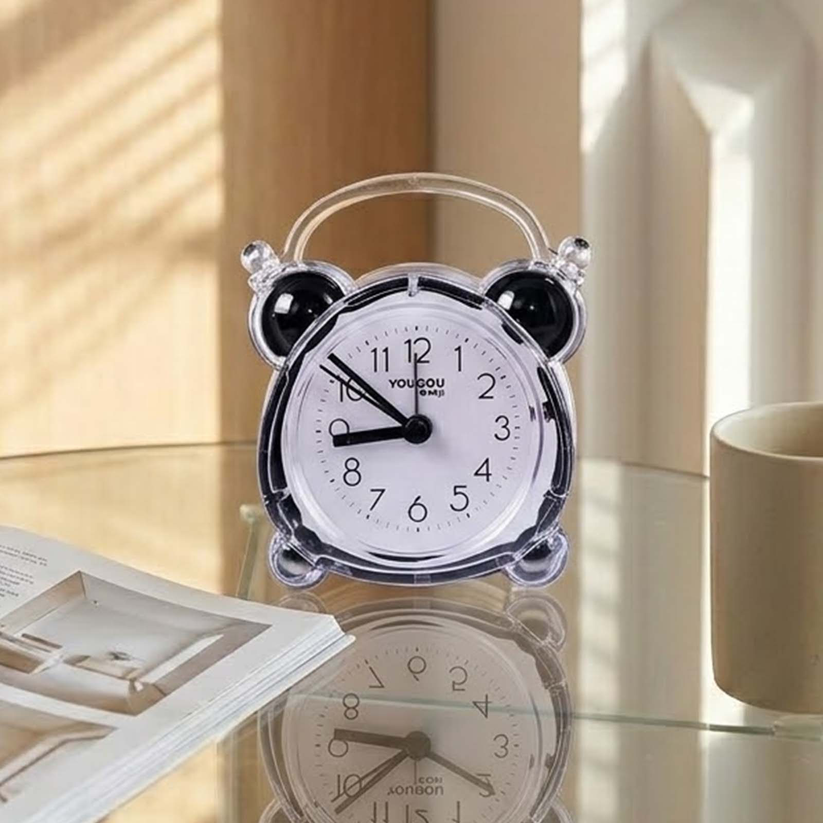Un despertador transparente con esfera negra sobre una mesa de cristal, reflejándose junto a un libro y una taza.