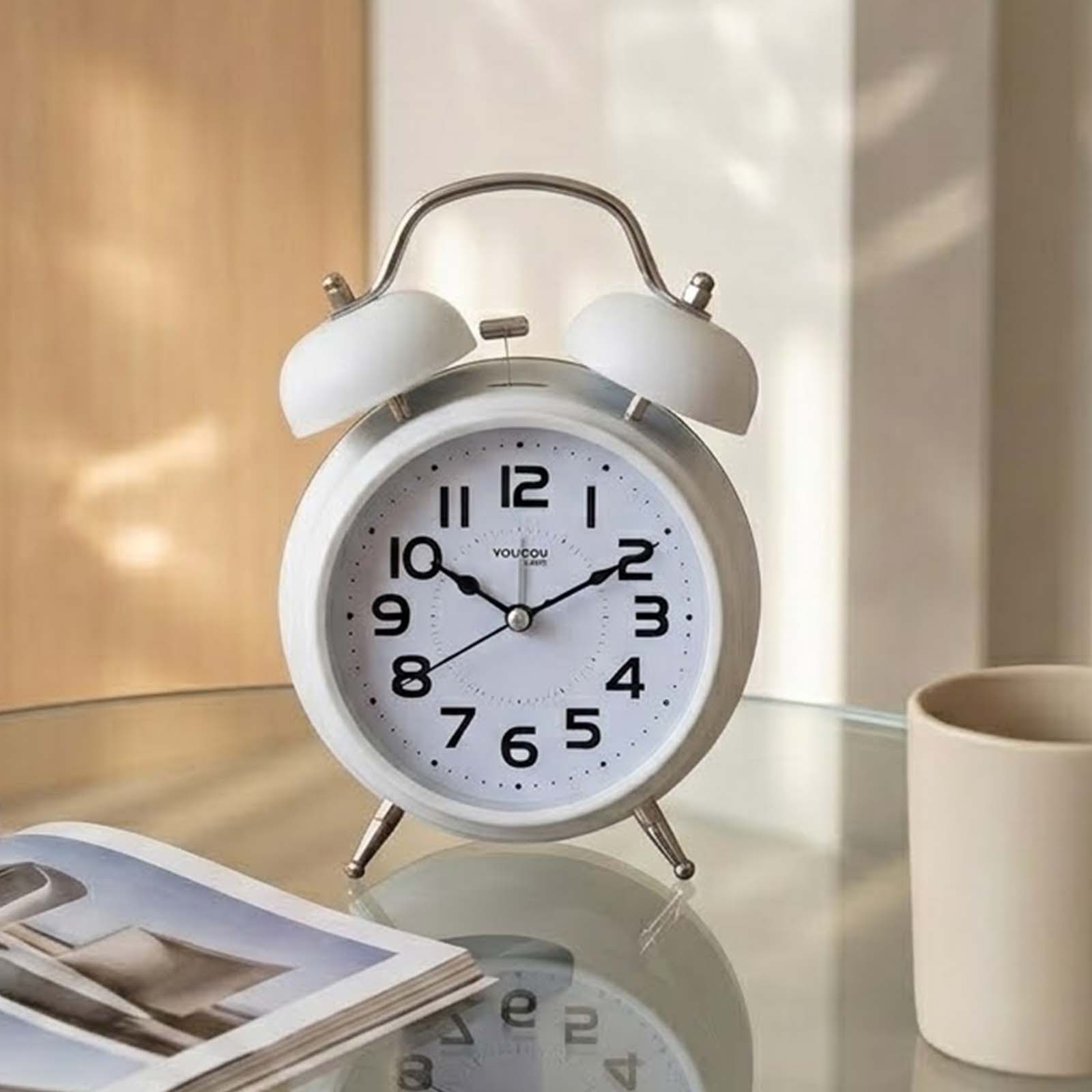 Un despertador retro blanco con números negros y patas de metal sobre una mesa de vidrio junto a una taza.