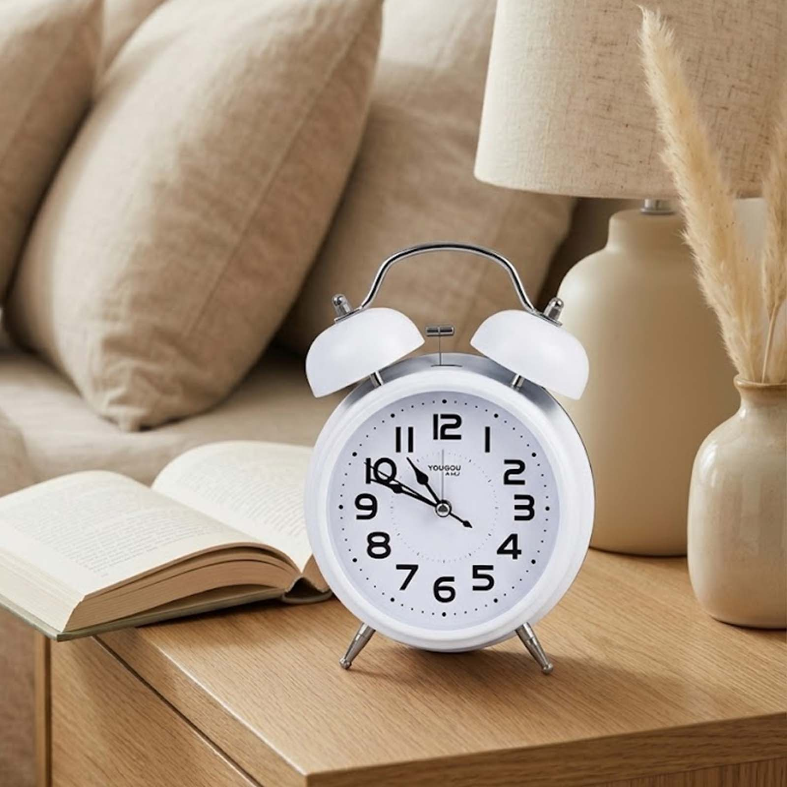 Un reloj despertador blanco sobre una mesita de madera junto a un libro abierto y una lámpara.