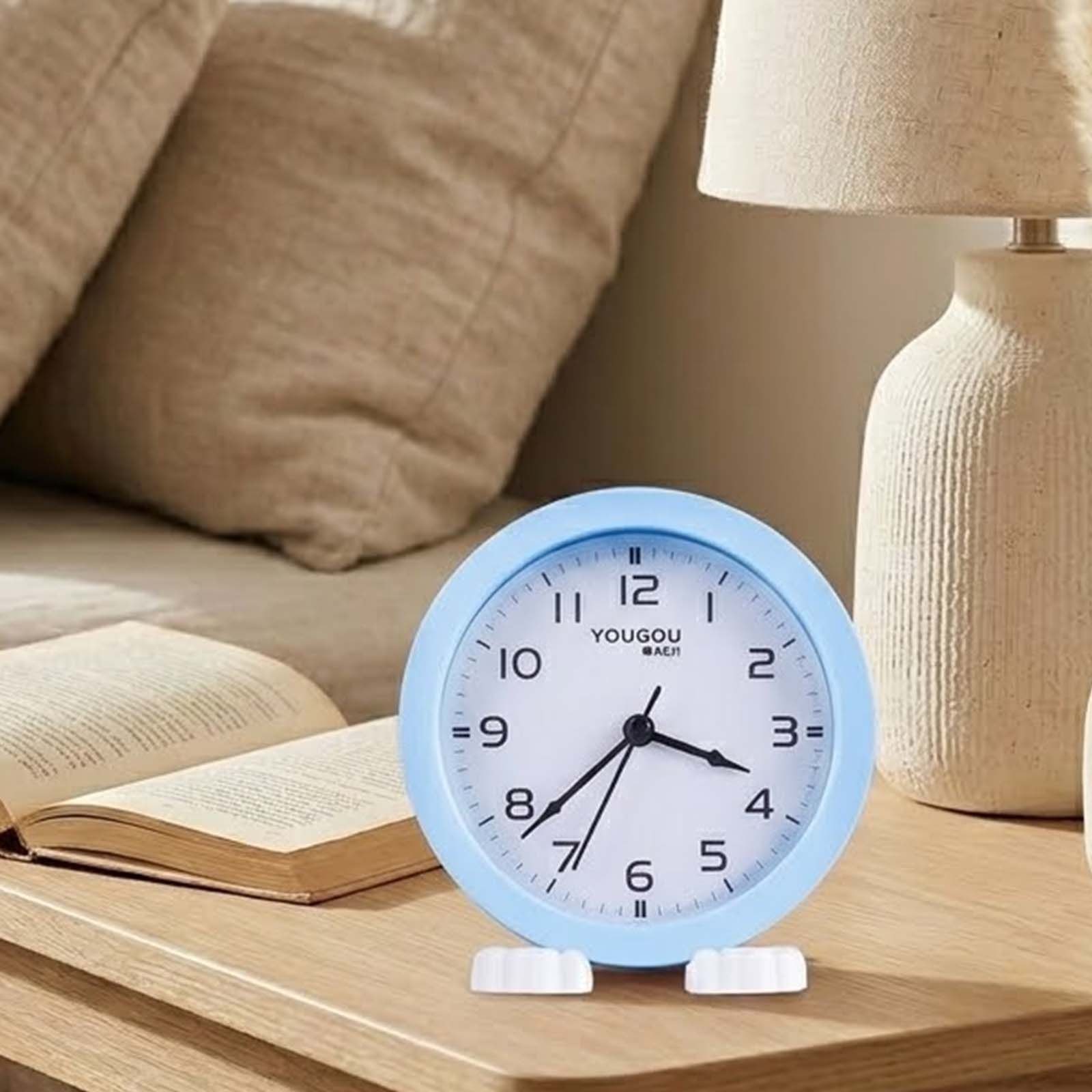 Un reloj despertador azul sobre una mesa de madera junto a un libro abierto y una lámpara.
