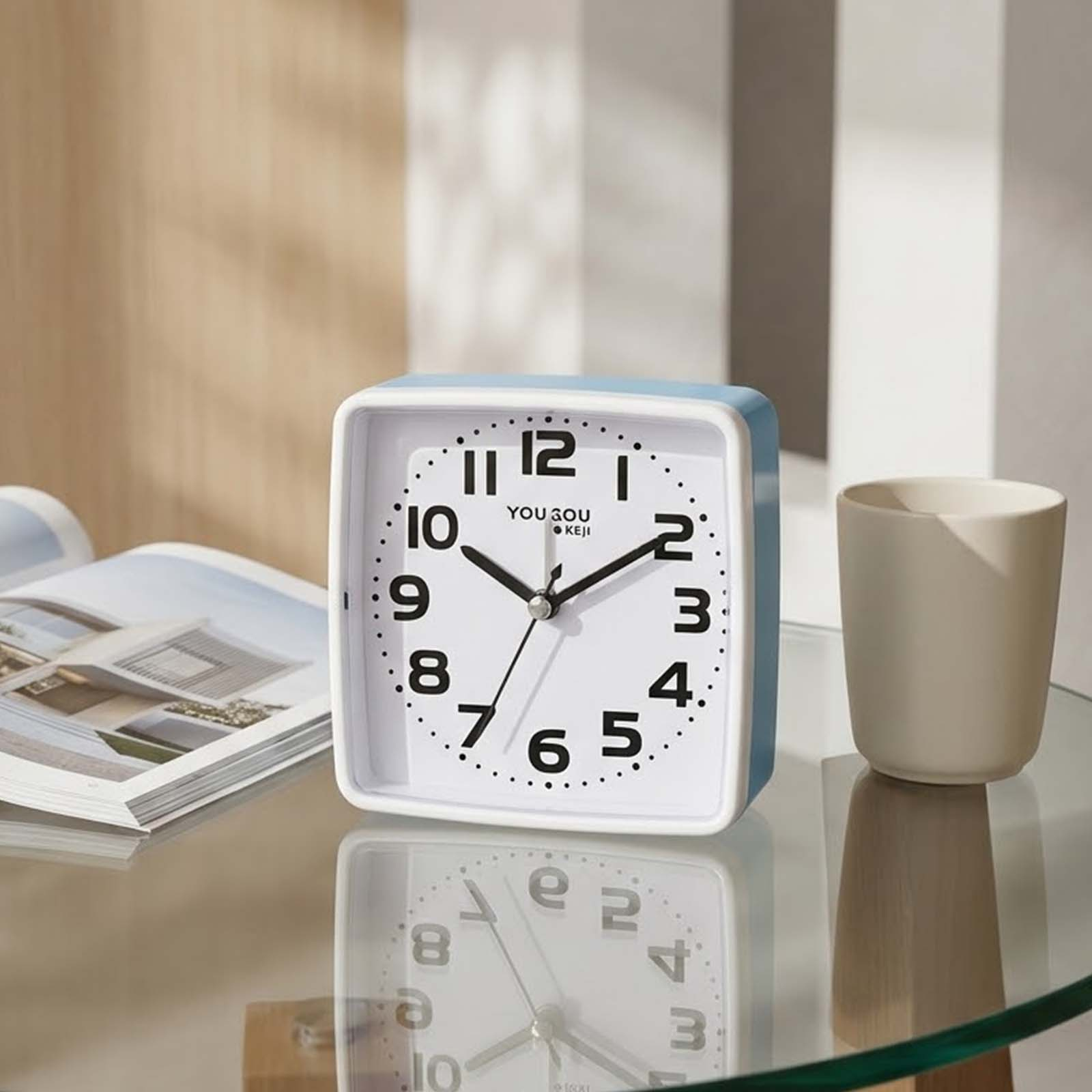 Un despertador cuadrado azul claro y blanco se encuentra en una mesa de vidrio junto a una revista abierta y una taza blanca.