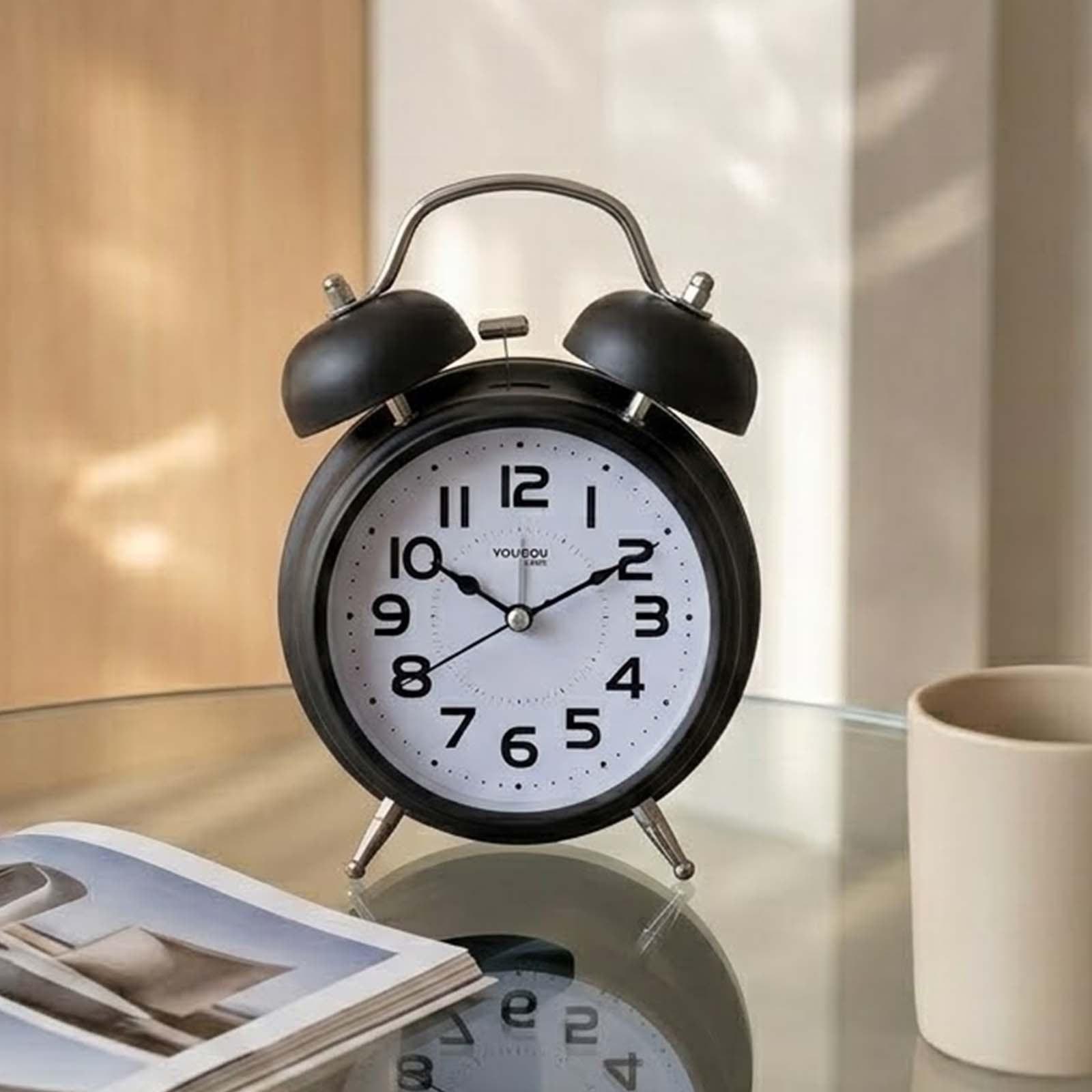 Un despertador retro negro con esfera blanca sobre una mesa de vidrio junto a una taza y una revista.