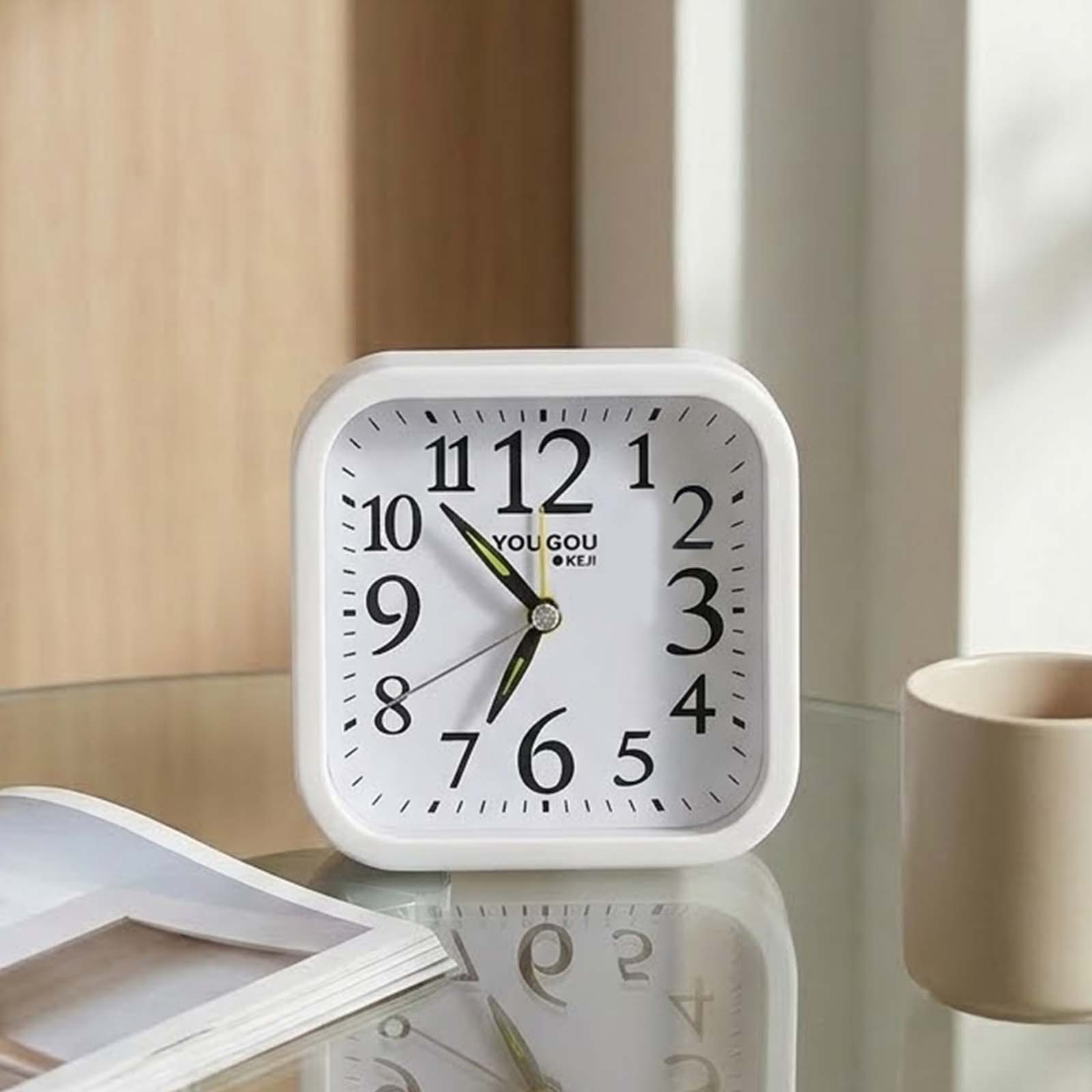 Un despertador cuadrado blanco en una mesa de cristal con taza y libro.