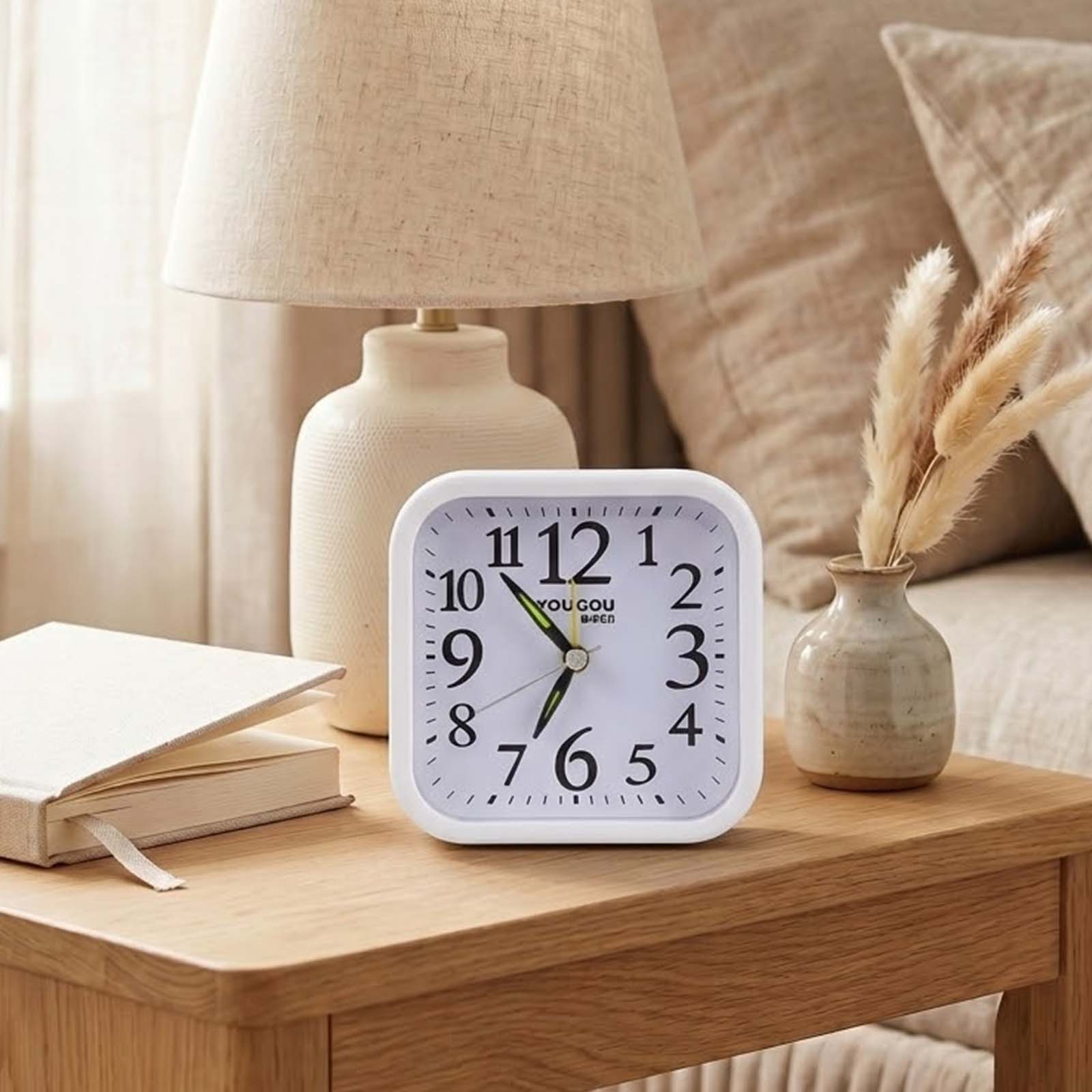 Un reloj cuadrado blanco sobre una mesa de madera junto a una lámpara, un libro y un pequeño jarrón con ramas secas.