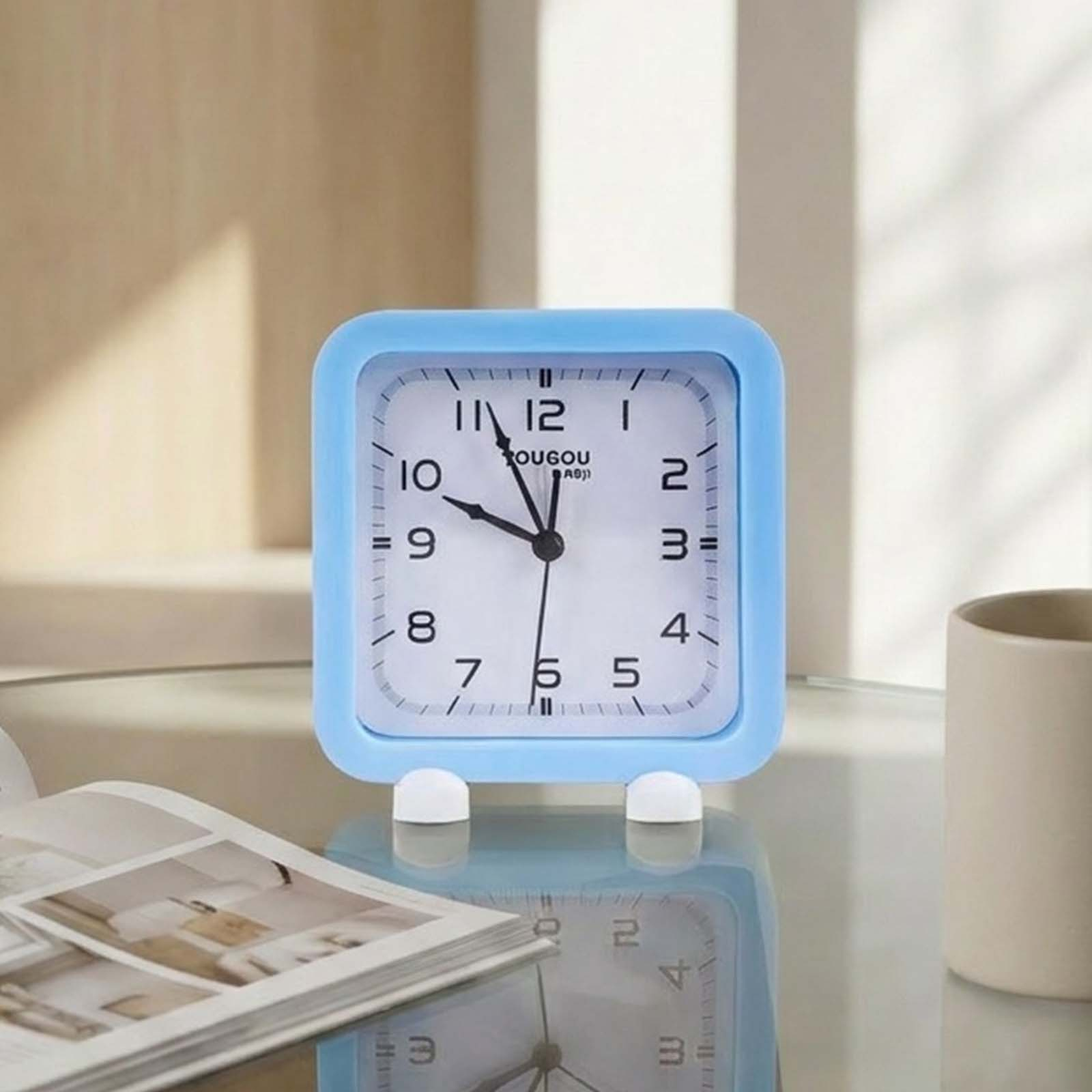 Un despertador cuadrado azul está sobre una mesa brillante junto a una revista y una taza.