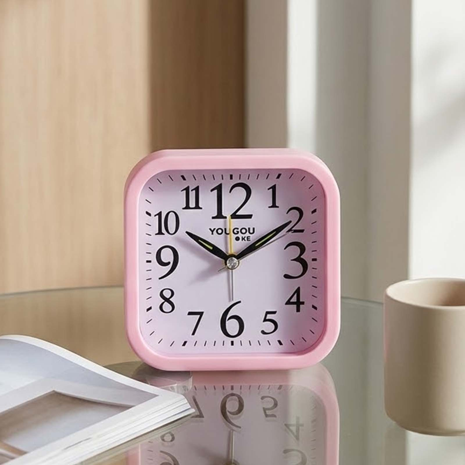 Despertador cuadrado rosa en mesa de cristal con taza y libro.