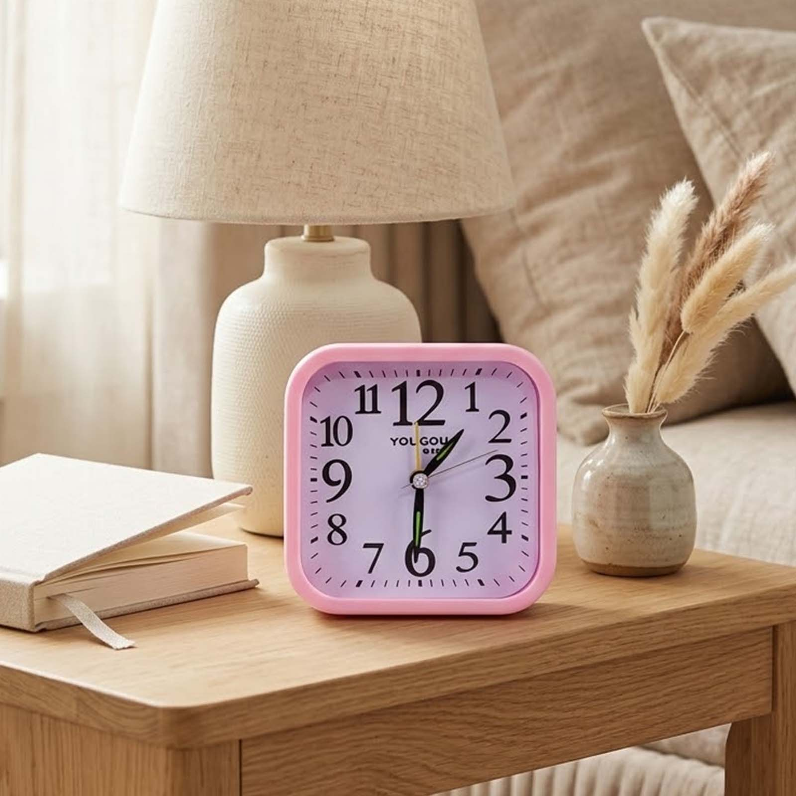 Un despertador cuadrado rosa sobre una mesa de madera junto a una lámpara, un libro y un jarrón pequeño.