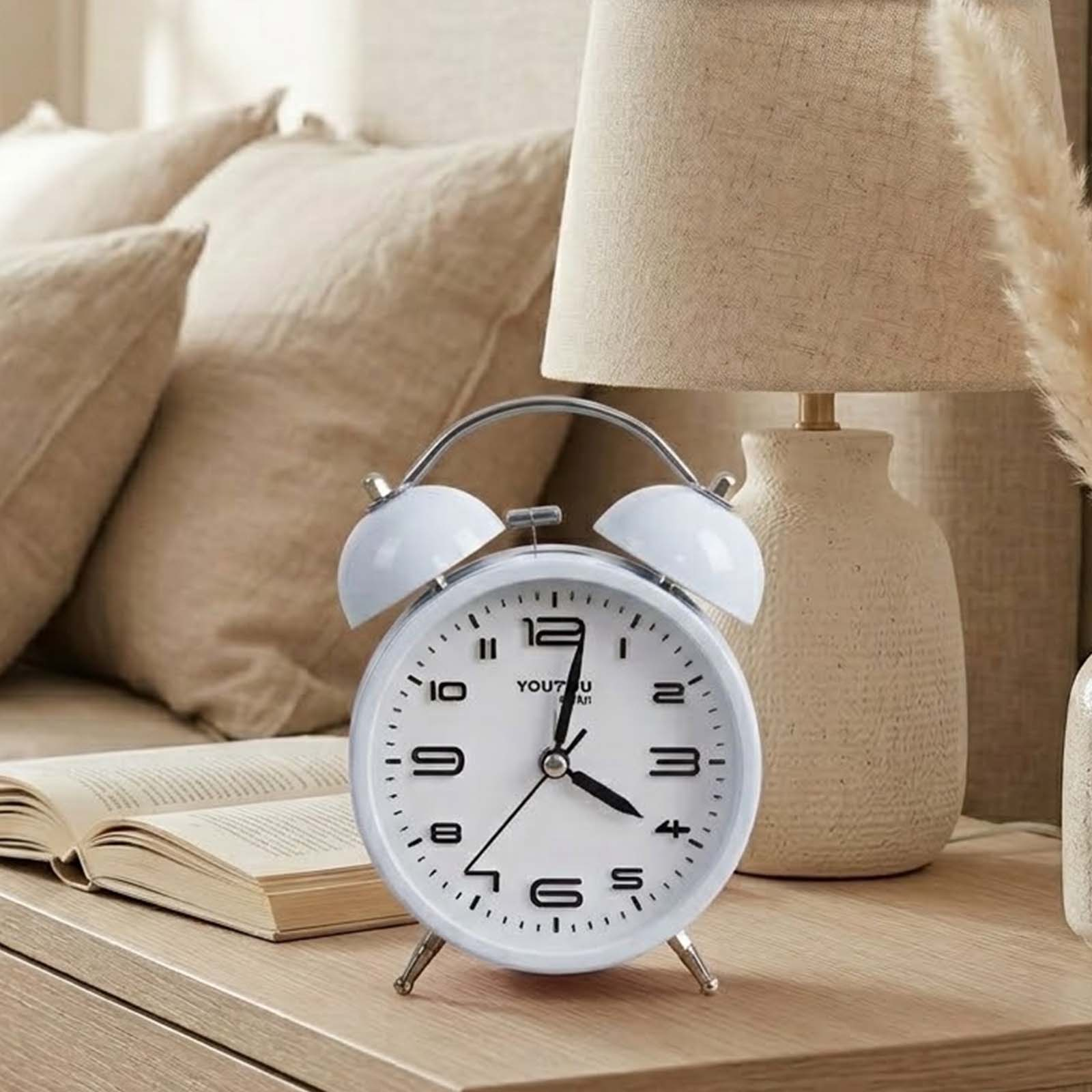 Un despertador blanco con doble campana sobre una mesita de noche, con un libro, lámpara y cojines.