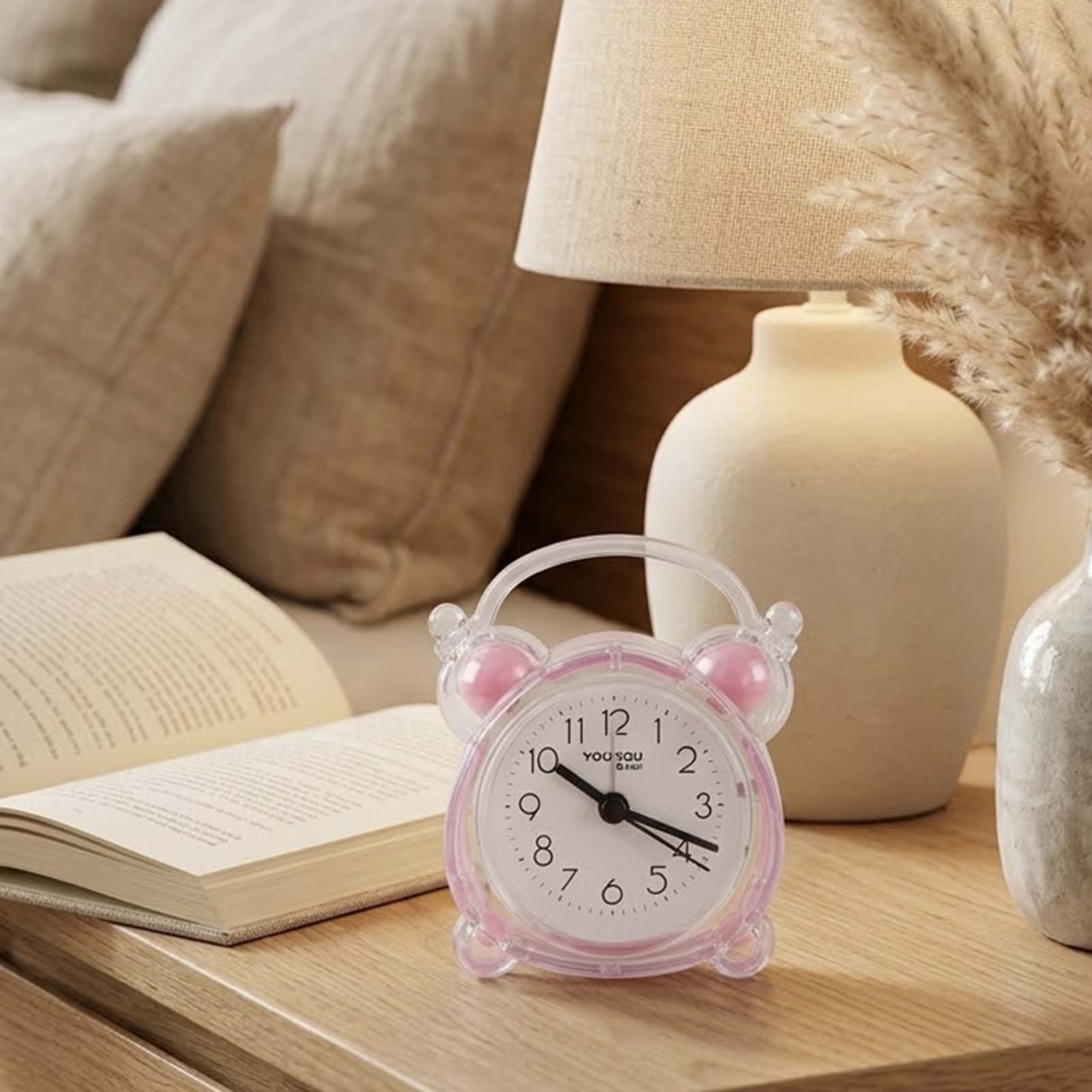 Un despertador de plástico rosa en una mesita de noche, con un libro abierto, lámpara y cojines.