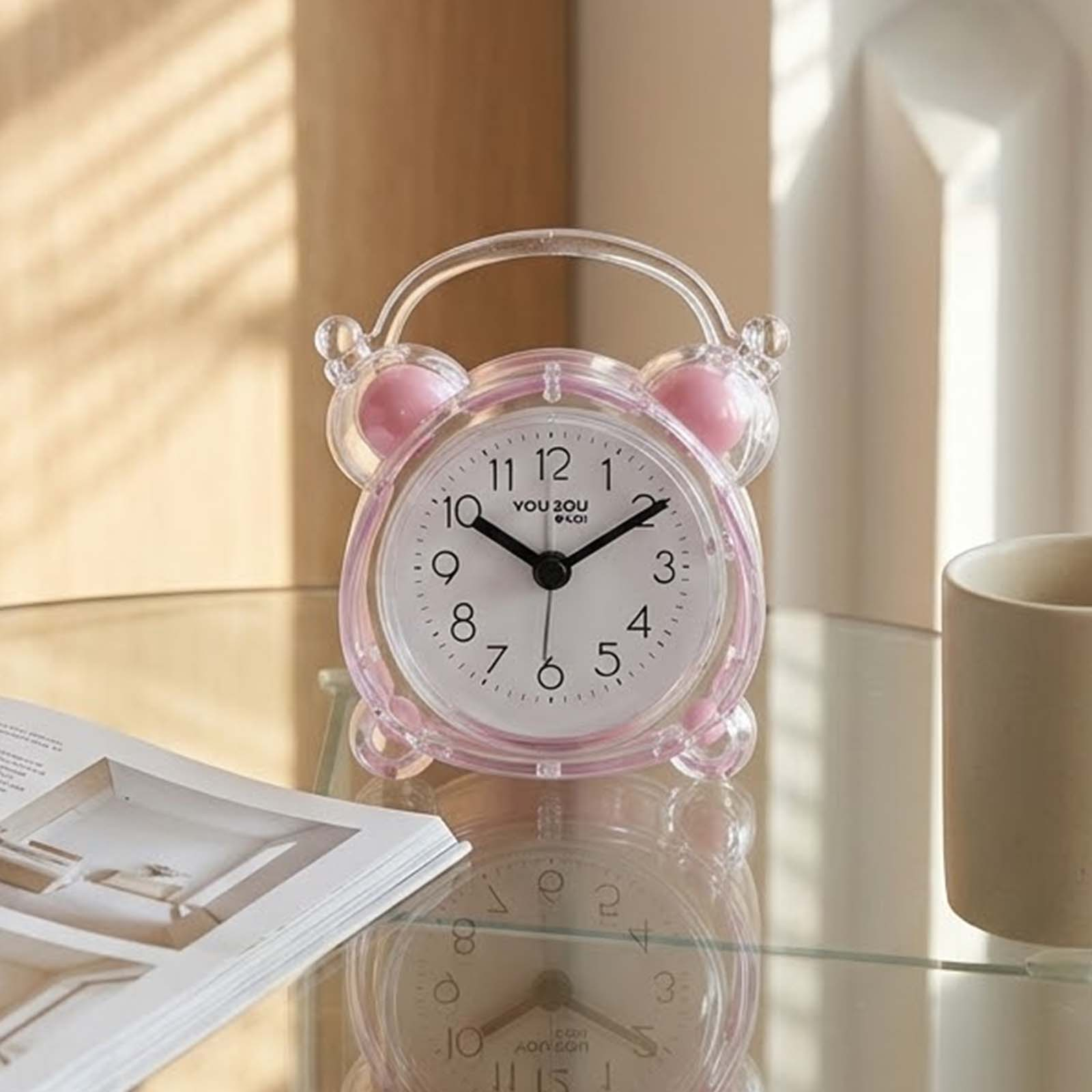 Un despertador transparente con detalles rosados descansa en una mesa de cristal junto a un libro y una taza.