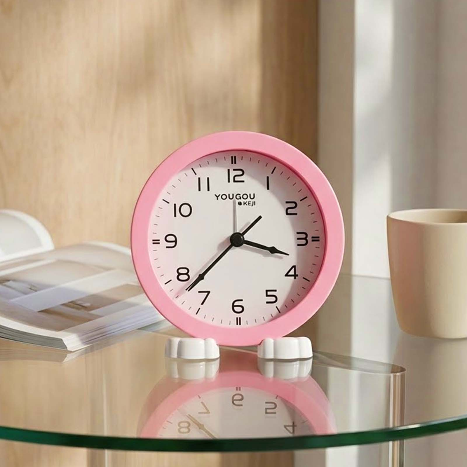 Un reloj de mesa analógico rosa con números y manecillas negras sobre una mesa de vidrio, con un libro abierto y una taza.