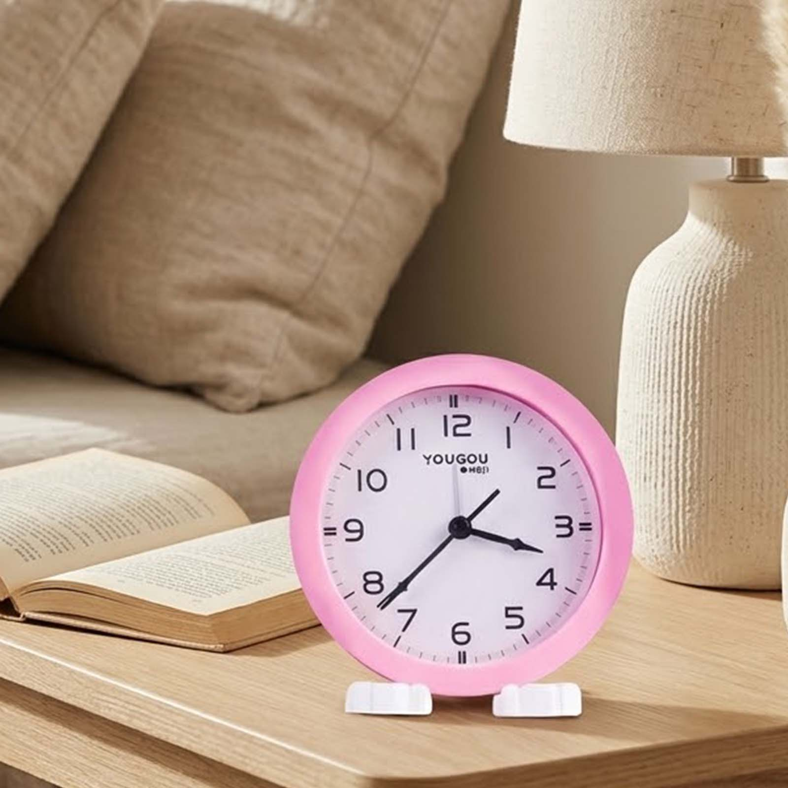 Un reloj de mesa analógico rosa se encuentra en una superficie de madera junto a un libro abierto y una lámpara.