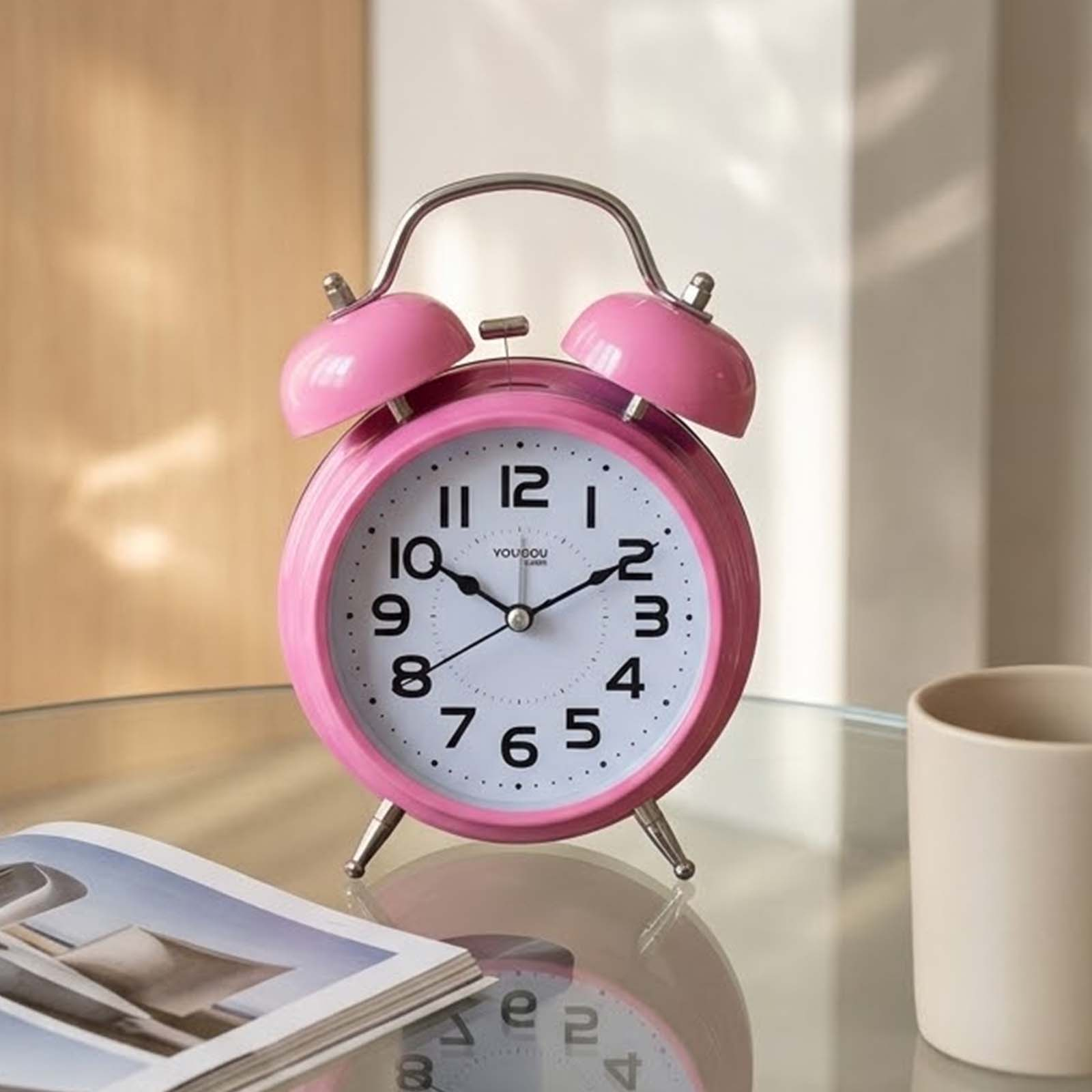 Un despertador rosa redondo con números blancos se encuentra en una mesa de cristal junto a una revista y una taza.