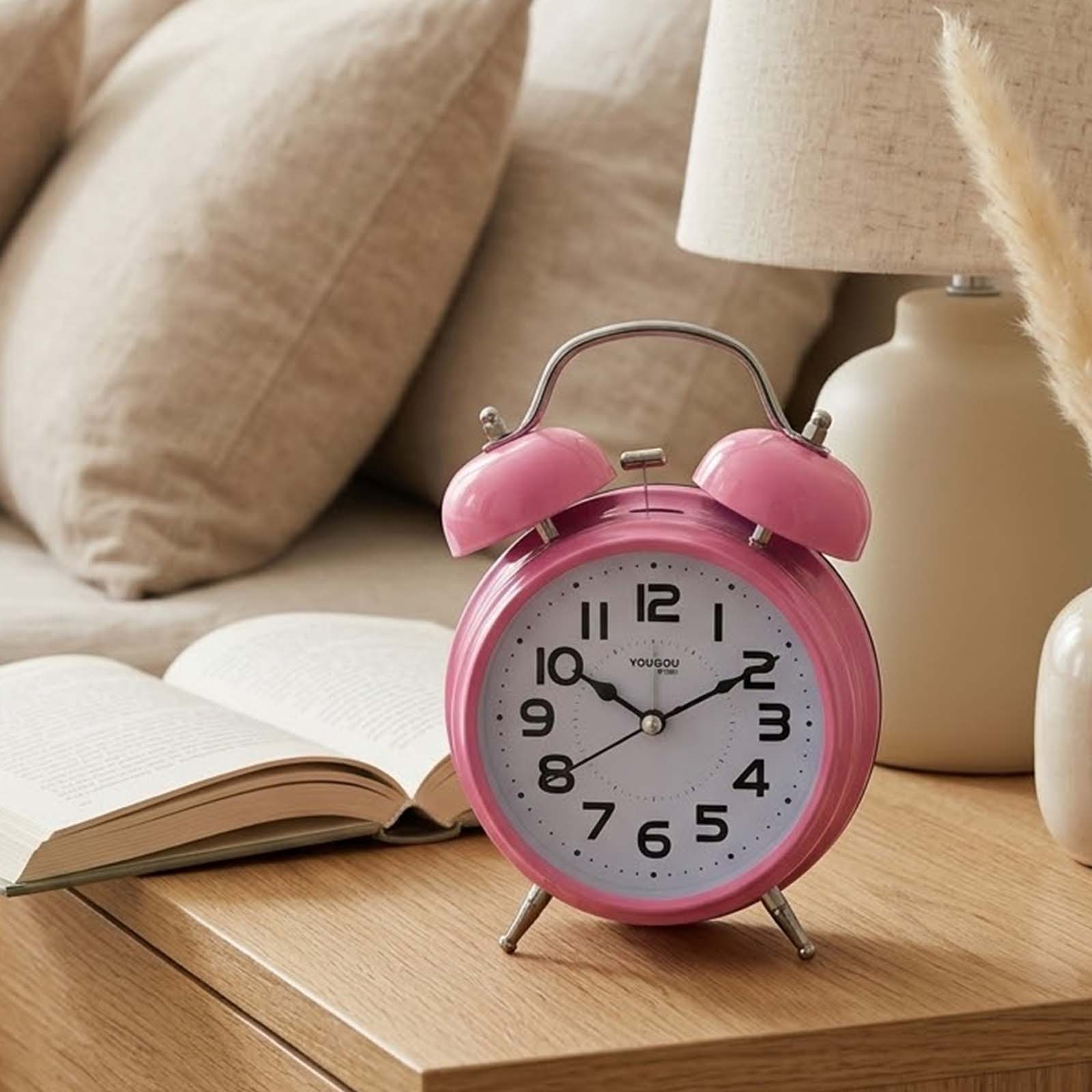 Un despertador rosa en una mesita de noche, con un libro abierto y una lámpara al fondo.