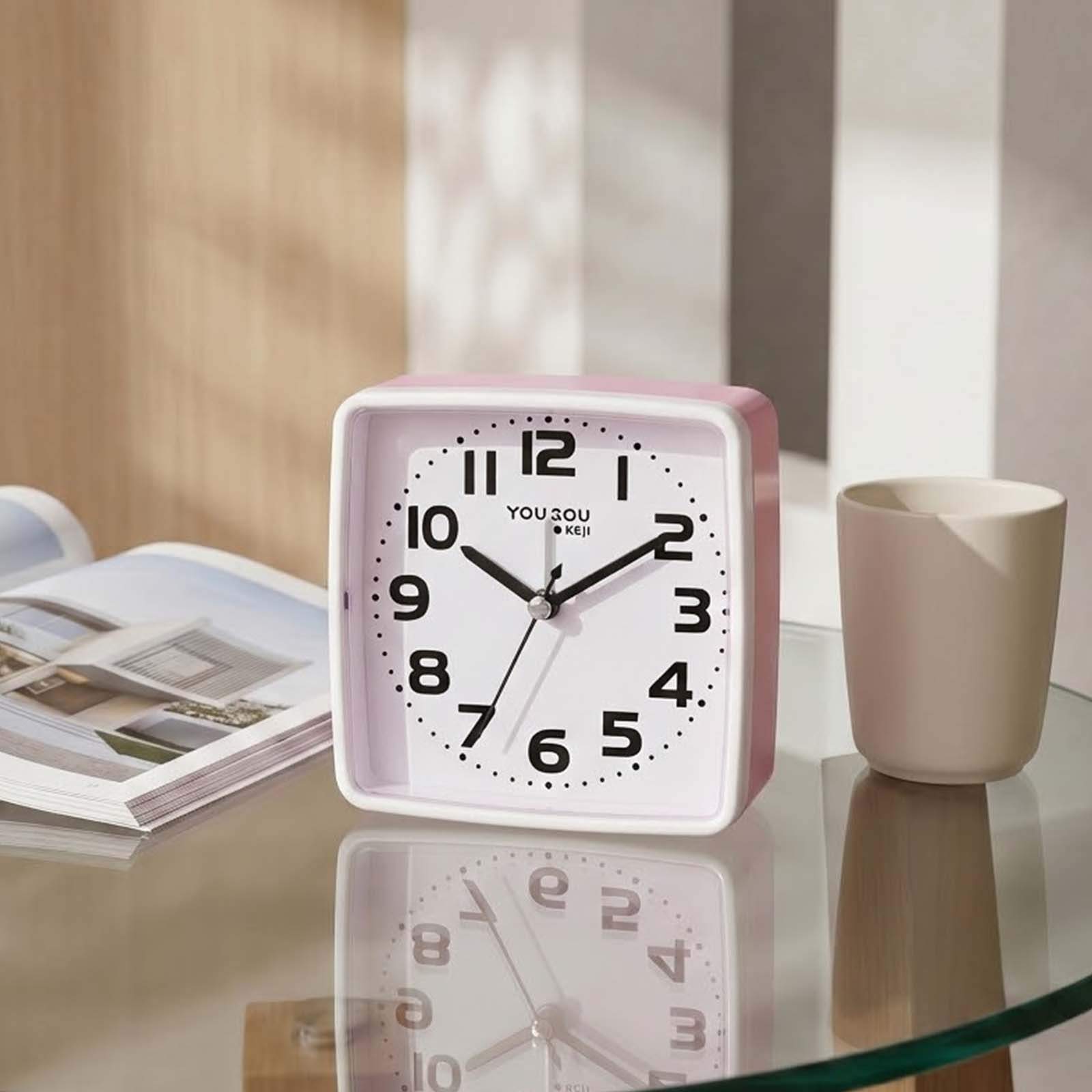 Un reloj de mesa cuadrado rosa con números negros se encuentra en una mesa de cristal junto a un libro y una taza.
