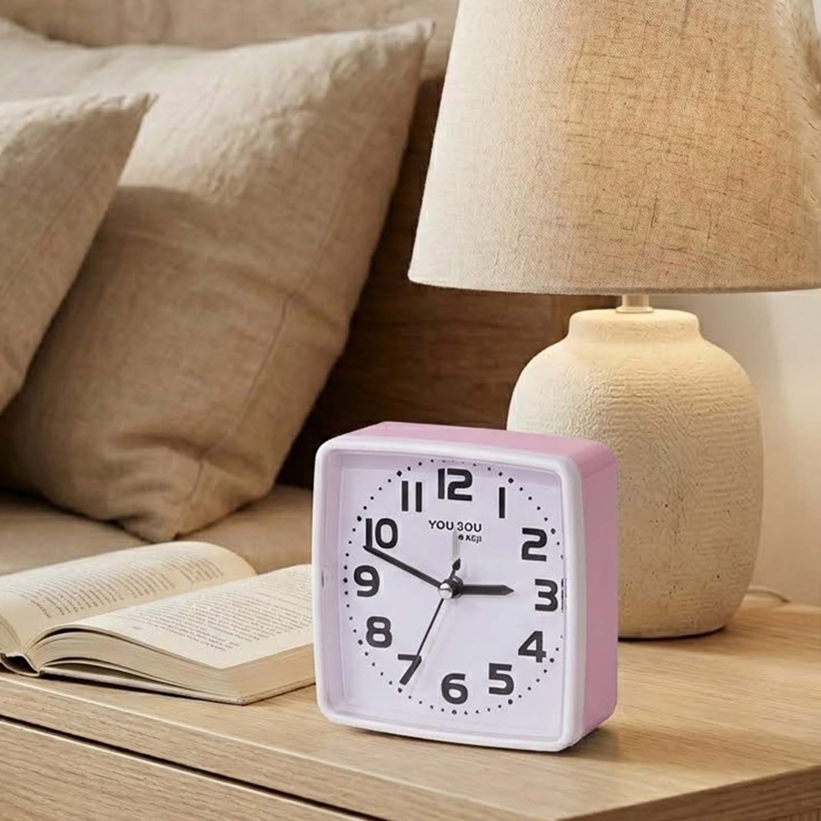 Un despertador rosa, un libro abierto y una lámpara de mesa están en una mesita de noche.
