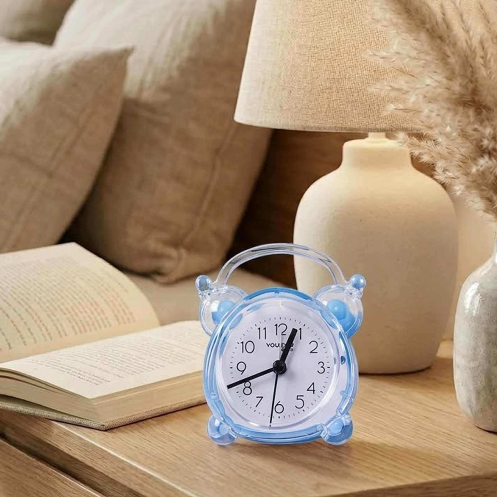 Un despertador azul sobre una mesa de madera junto a un libro abierto y una lámpara, con cojines de fondo.