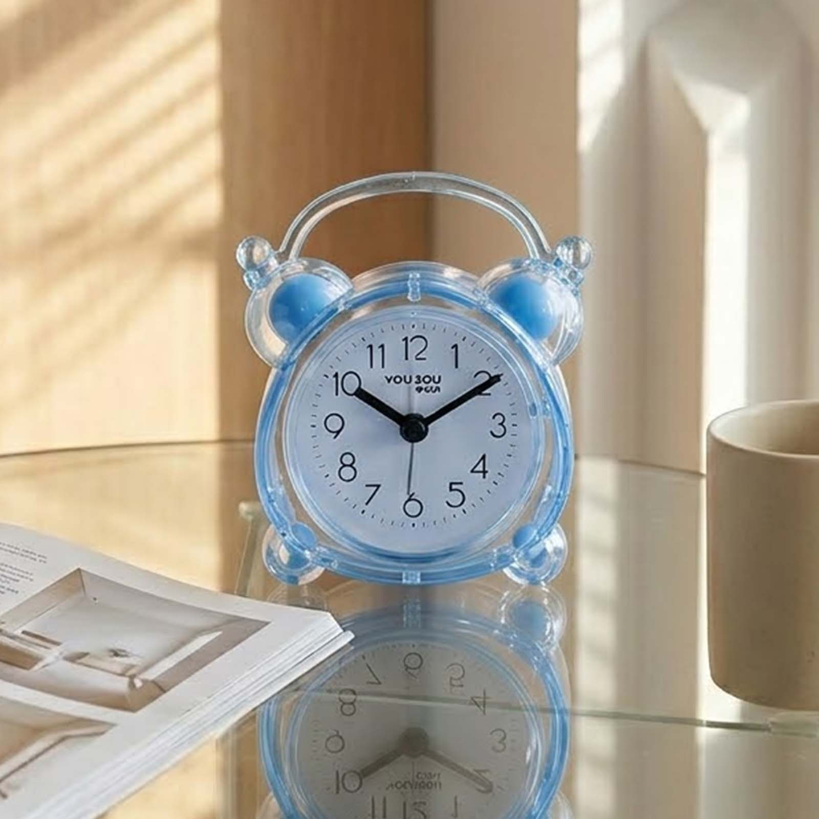 Un despertador transparente azul se encuentra sobre una superficie de vidrio junto a un libro y una taza.