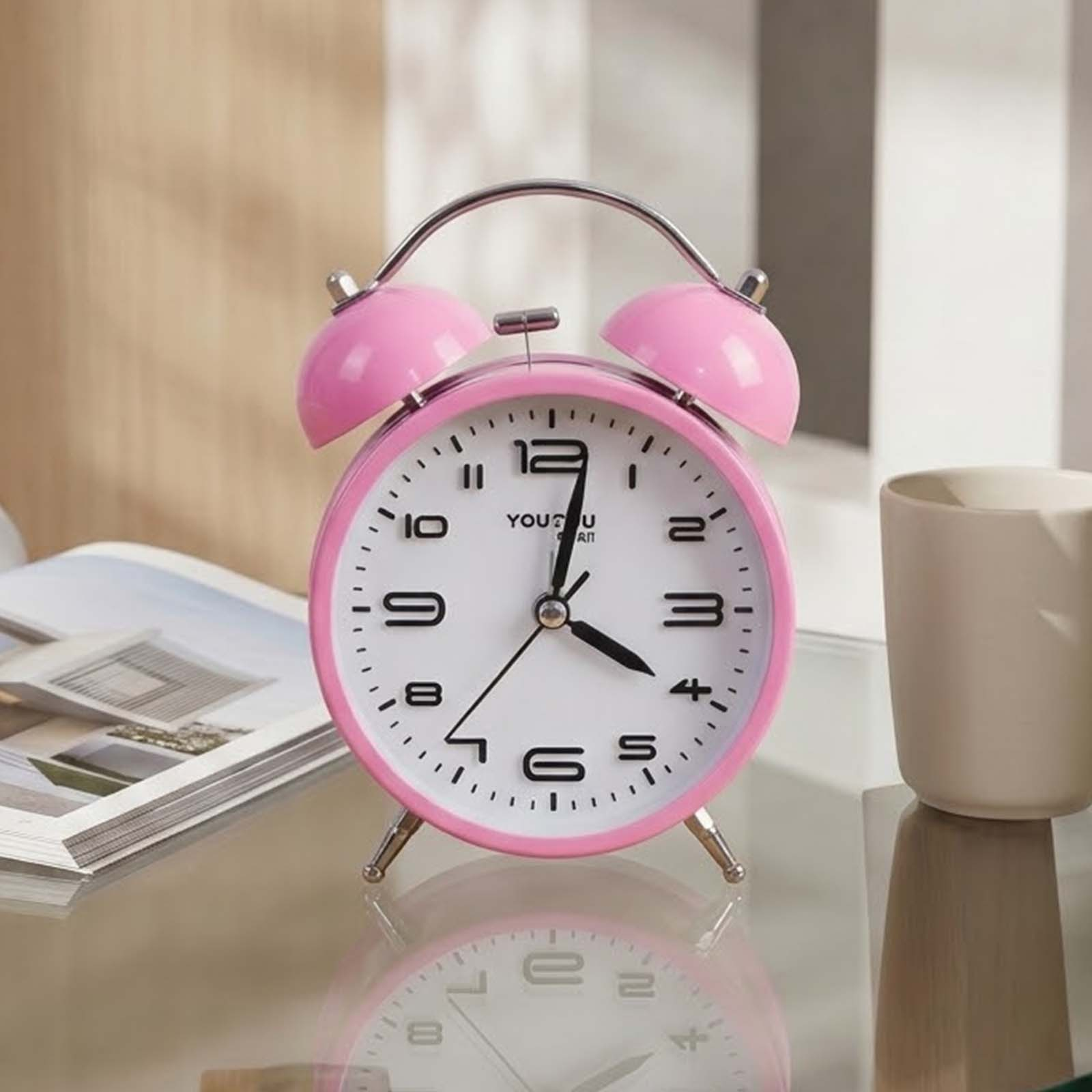 Un despertador retro rosa sobre una mesa de vidrio al lado de una taza y una revista.