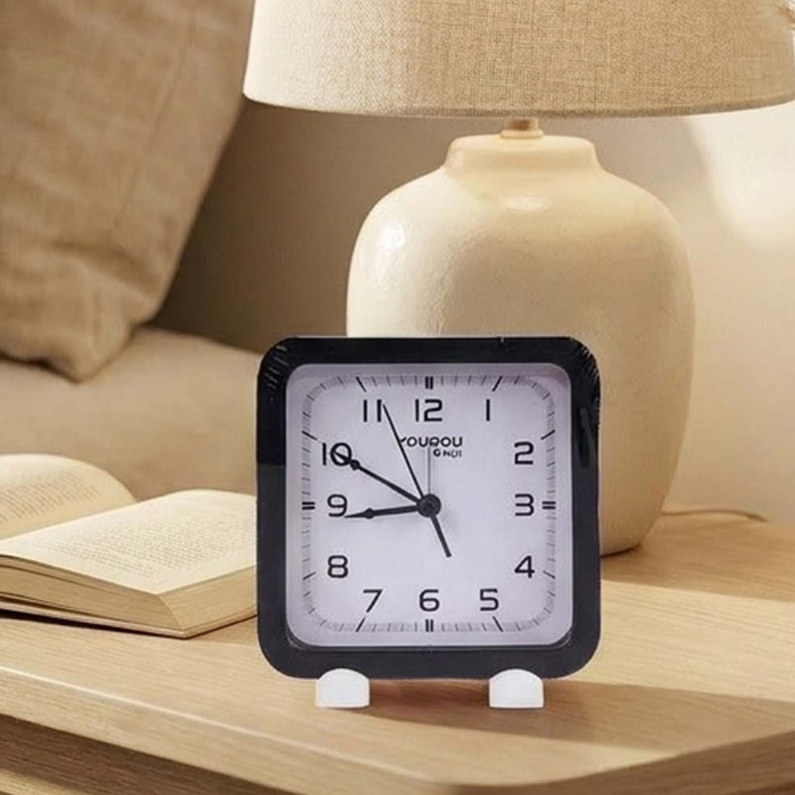 Un reloj despertador cuadrado con marco negro sobre una mesa de madera, con una lámpara y un libro al fondo.