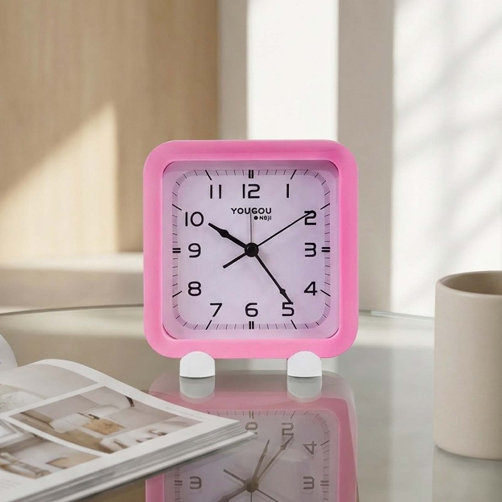 Despertador cuadrado rosa en una mesa de cristal con un libro y una taza.