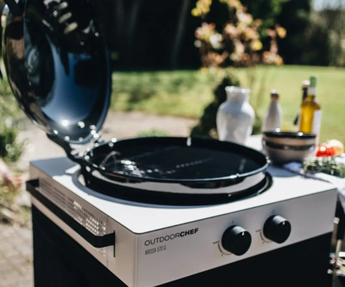 Açık havada, kapağı açık bir Outdoorchef gazlı mangal, bulanık bir arka plan önünde duruyor.