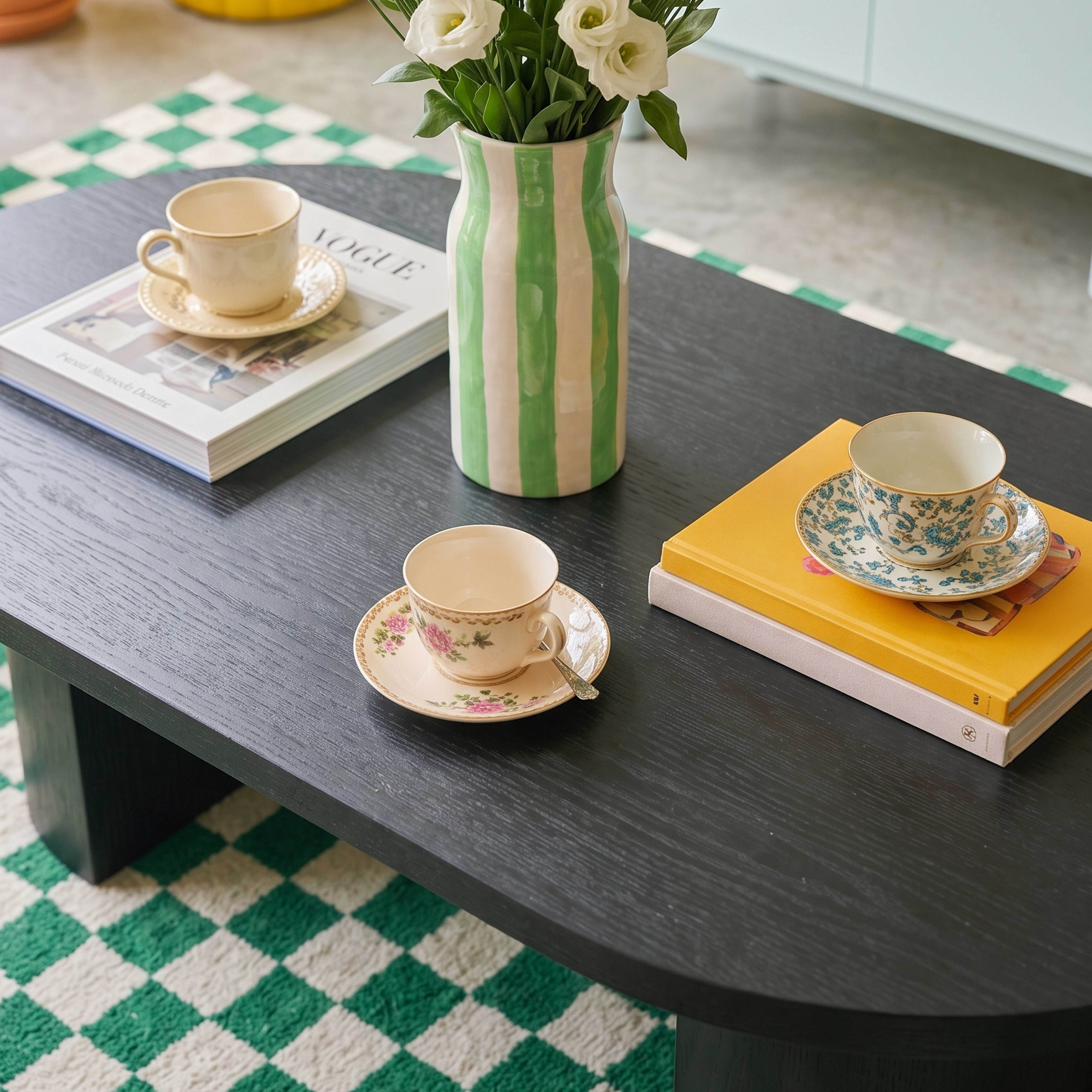 Ein schwarzer Couchtisch mit drei Teetassen, zwei Büchern und einer grün-weiß gestreiften Vase mit Blumen.