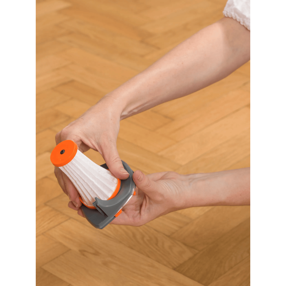Manos sosteniendo un filtro de aspiradora blanco y naranja sobre suelo de madera.