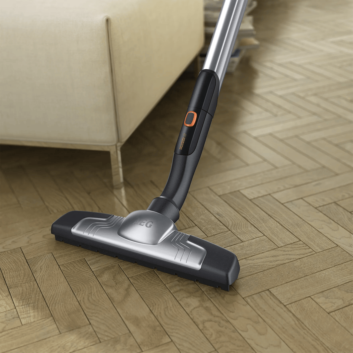 Eine silber-schwarze Staubsaugerdüse reinigt einen Fischgräten-Holzboden nahe einem beigen Sofa.