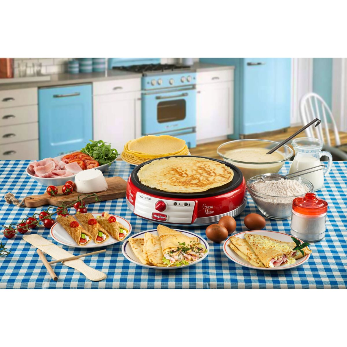 Una piastra per crepes rossa su un tavolo a quadretti blu, circondata da ciotole di ingredienti, uova e crepes cotte.